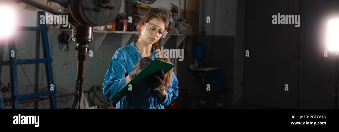 A young beautiful woman auto mechanic with a tablet in his hands is standing at a service station. makes an inspection of the car and notes the cause Stock Photo