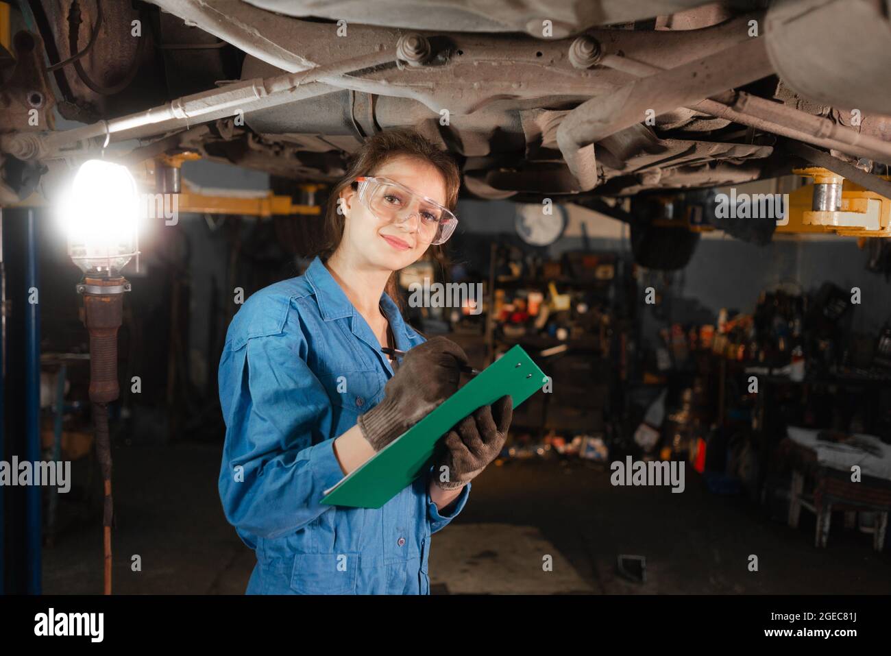 young beautiful woman car mechanic makes an inspection of the chassis of the car. stands in goggles and overalls with a tablet in his hands. Copy spac Stock Photo