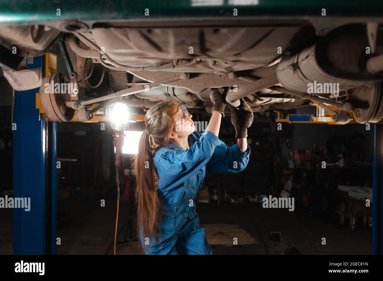 Young beautiful woman car mechanic dressed in protective goggles and working overalls makes an inspection of the chassis of the car. Copy space. Stock Photo