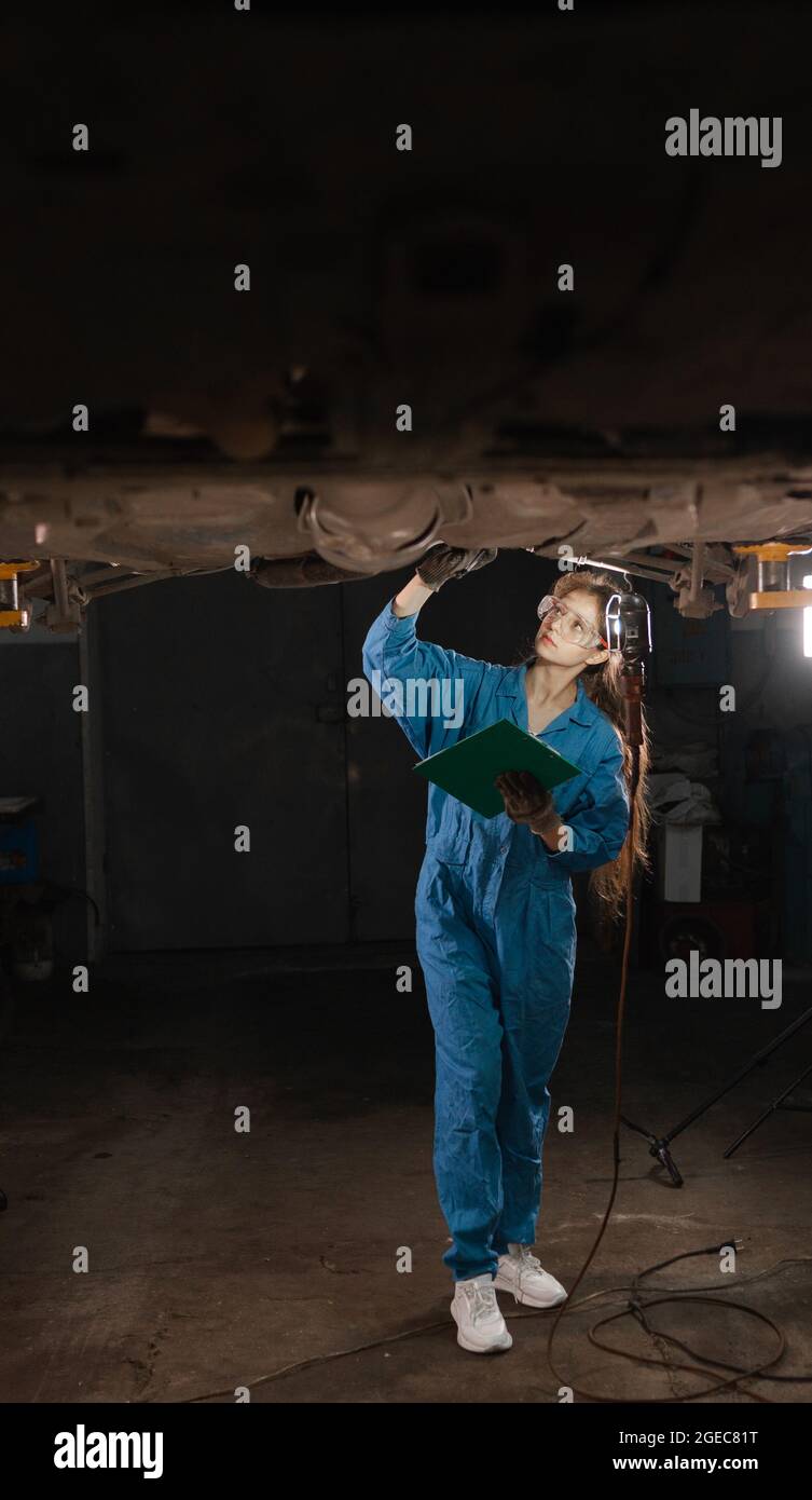 A beautiful lady car mechanic dressed in overalls and safety glasses stands under the raised car. makes an inspection of the undercarriage and looks f Stock Photo