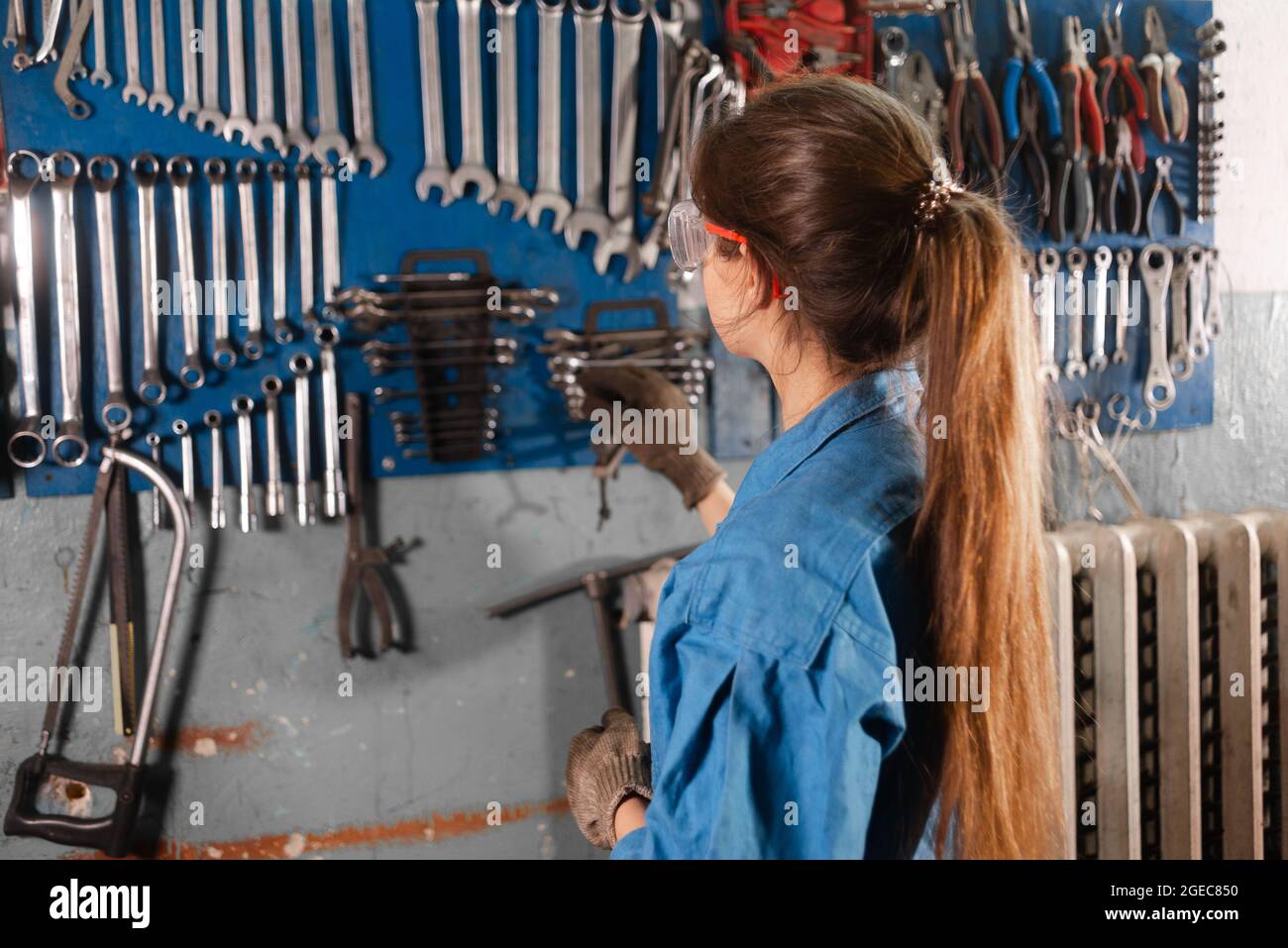 A young woman mechanic stands in the garage near the stand with the keys and makes a choice. Stock Photo