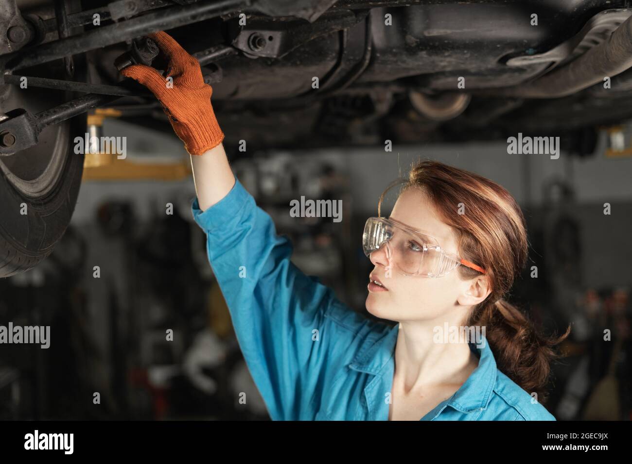 Young Cavsian woman auto mechanic at a car service station stands under a raised car and inspects the chassis. dressed in a blue overalls. Stock Photo