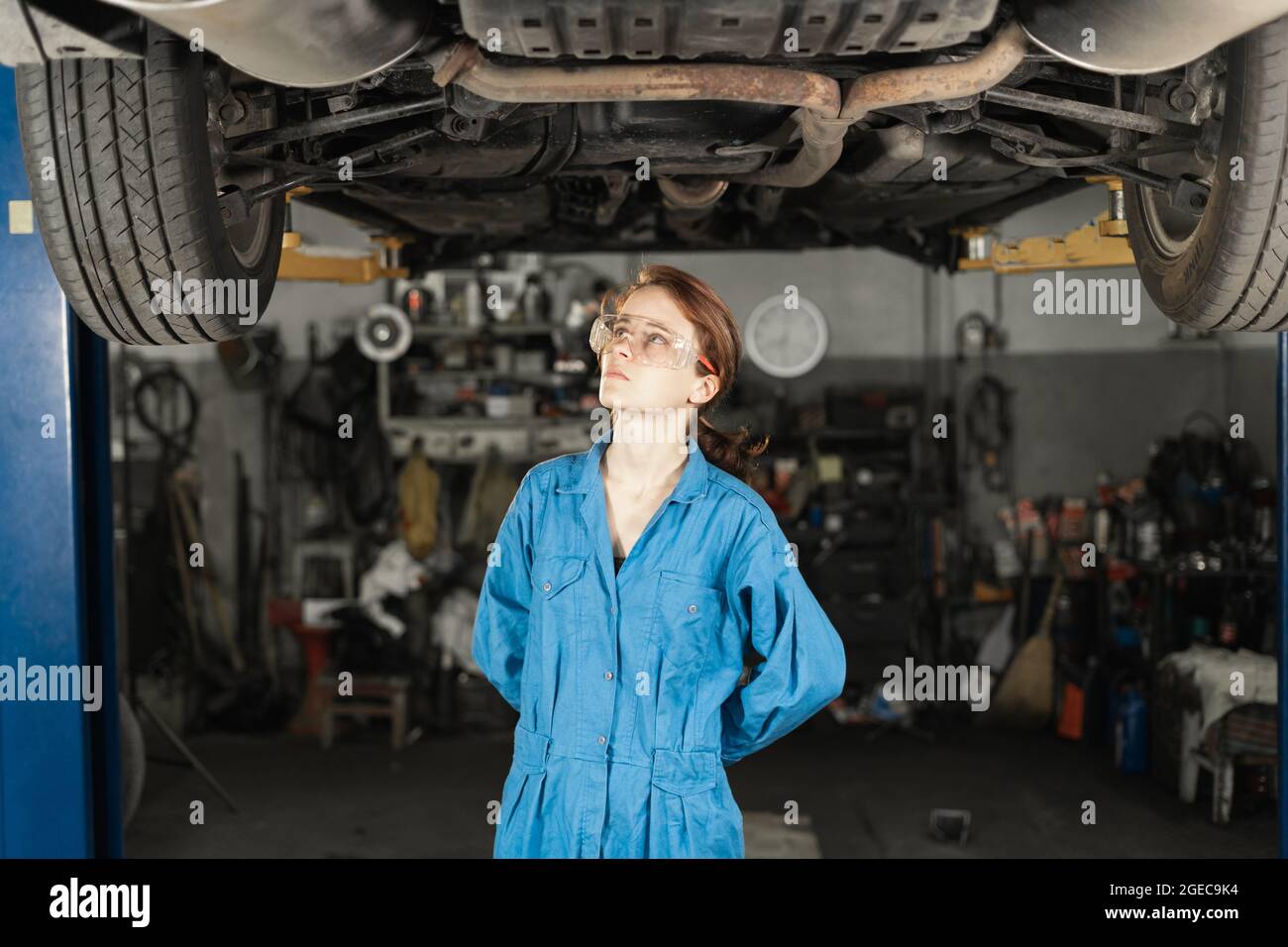 Auto workshop and a woman mechanic with glasses stands under the car in the garage. Inspection of the chassis and suspension of a car by a mechanic. G Stock Photo