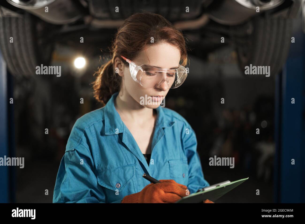 A woman mechanic stands against the background of a raised car. holds the tablet in his hands and records the damage. Marks with a pen. Copy space. 4k Stock Photo