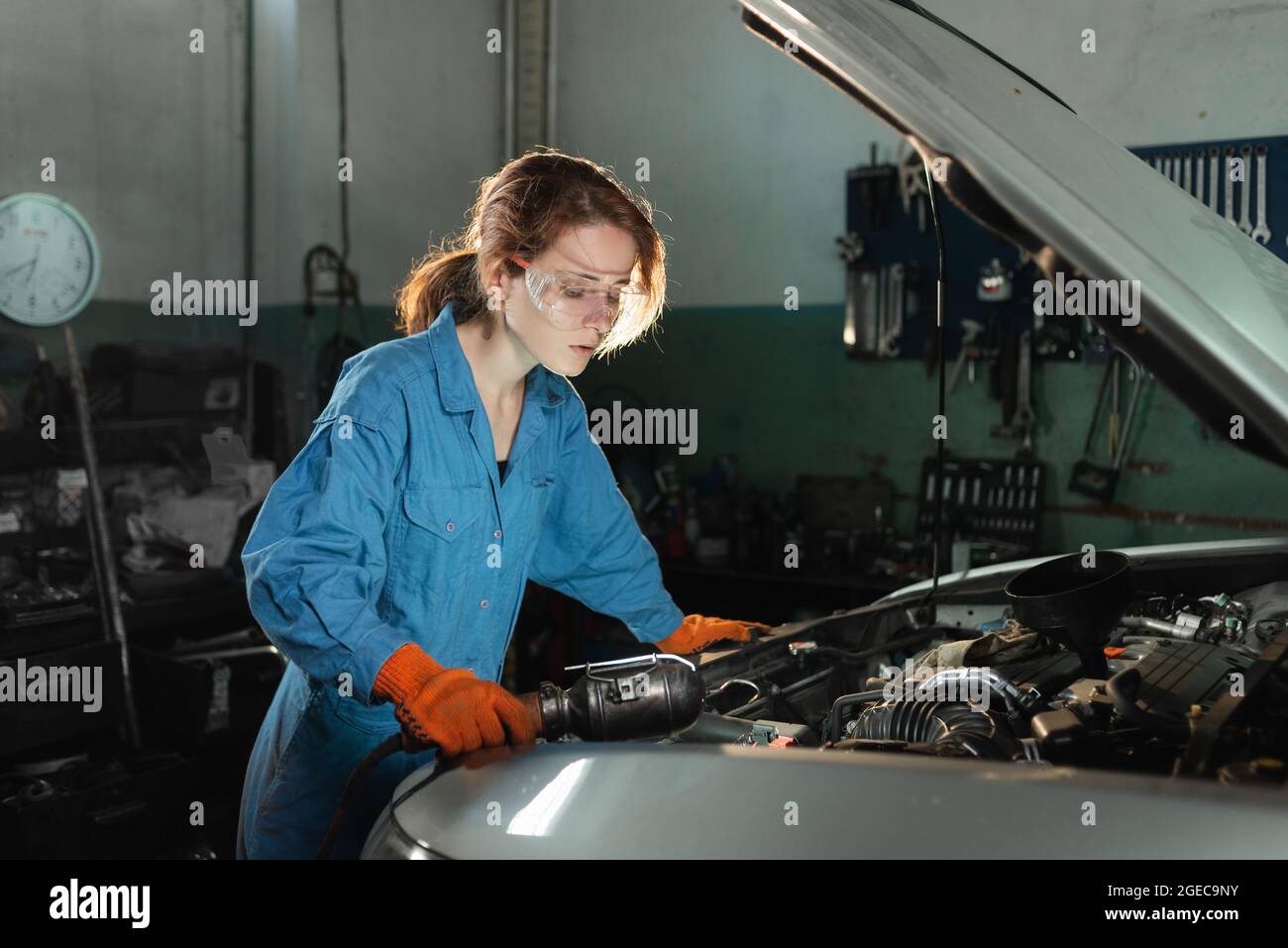 A young beautiful woman car mechanic is standing near the open hood of a car and makes a technical inspection at a service station. Stock Photo