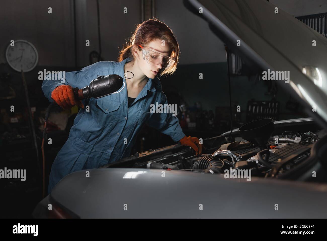 A woman mechanic makes an inspection of a car engine at a service station dressed in work uniforms overalls. Stock Photo