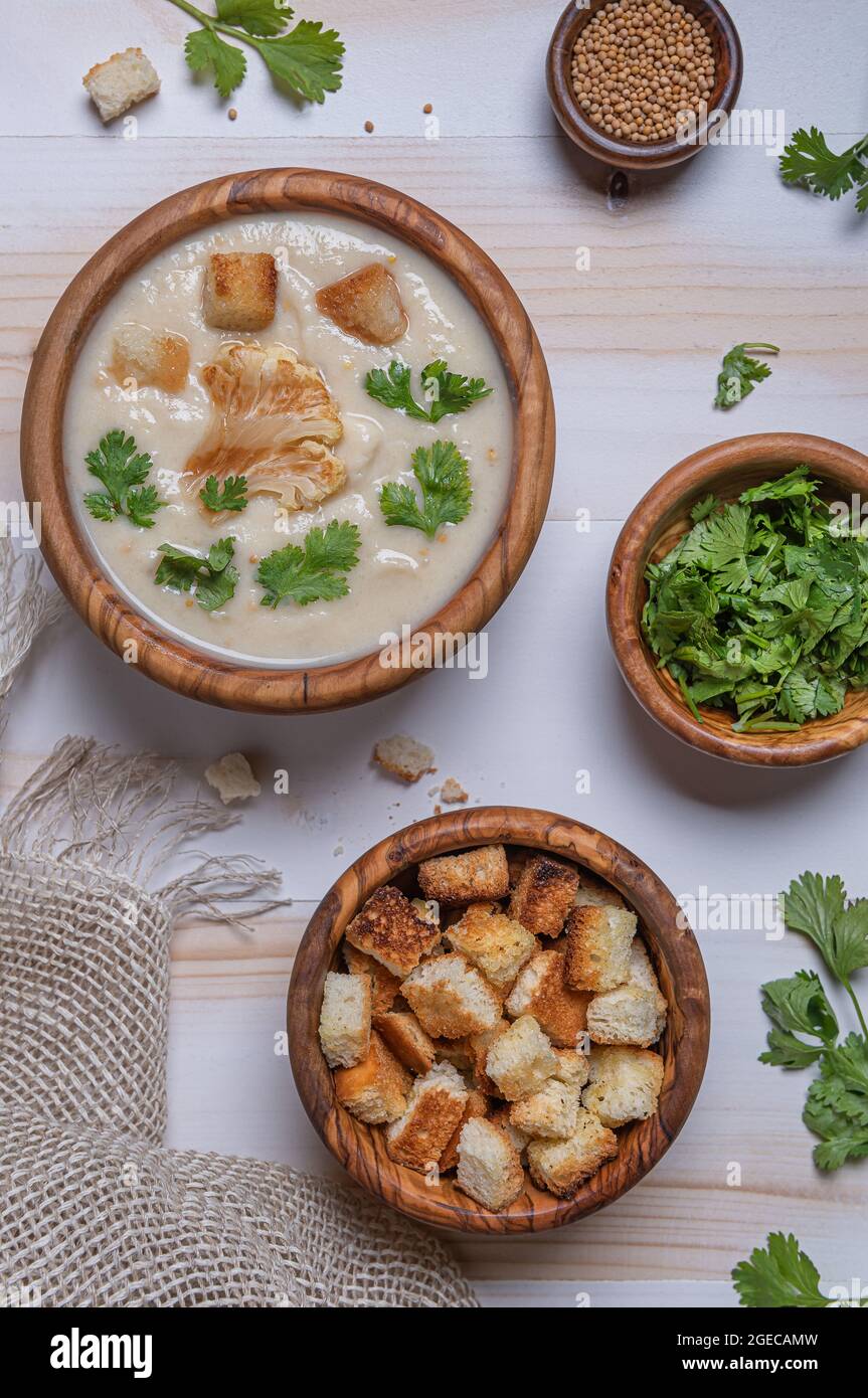 Flat lay with freshly made white cauliflower cream soup, fresh green herbs and croutons in wooden bowls on wooden white table. Linen napkin, mustard s Stock Photo