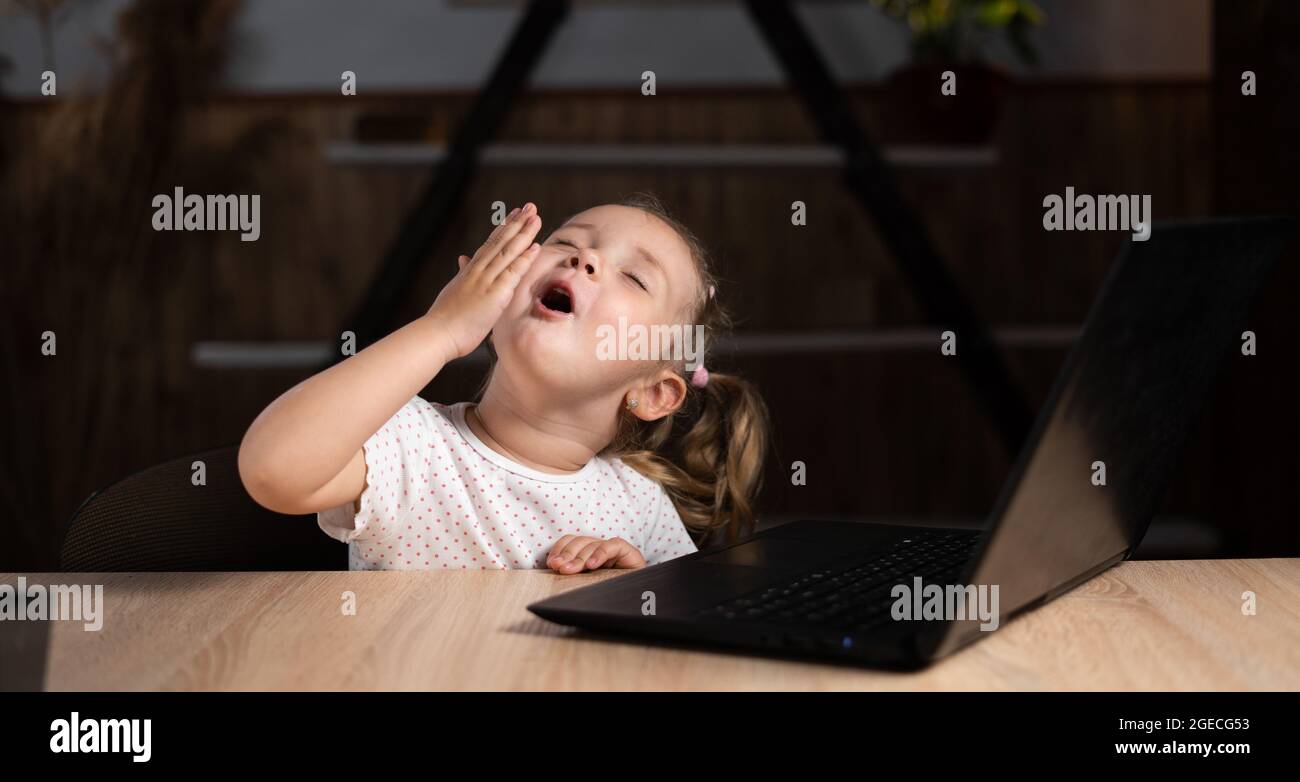 A preschool girl in the evening sits at a table with a laptop and yawns. Stock Photo