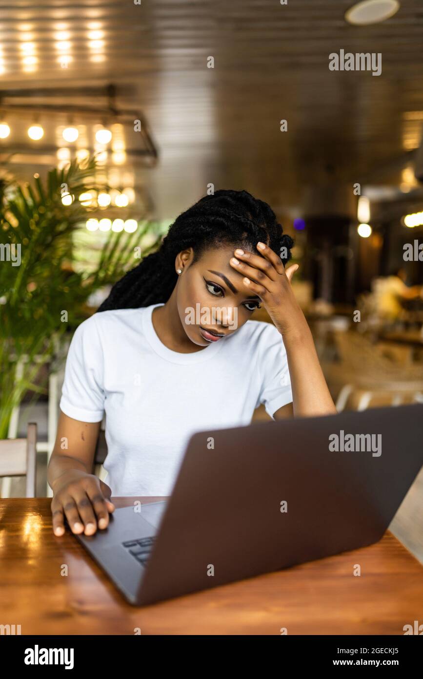 Pretty African woman sitting at table near laptop and looking at camera with tired face expression. Stock Photo