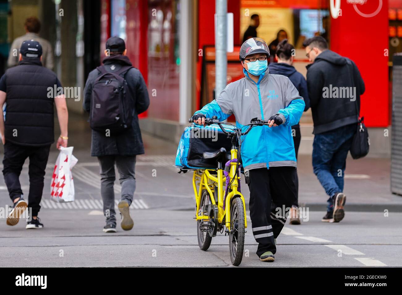 Melbourne, Australia, 21 June, 2020. MELBOURNE, AUSTRALIA - June 21: A food delivery rider is seen wearing a facemask as he walks his bicycle across an intersection in Melbourne as restrictions are being tightened in Victoria during COVID 19 on 21 June, 2020 in Melbourne, Australia. After a concerning spike in new coronavirus infections in metropolitan Melbourne which is once again sparking fears of a second wave, the Premier Daniel Andrews announced yesterday (20 June) that restrictions on the number of visitors in private homes will be reduced to ten to five. Victoria recorded 19 new cases o Stock Photo