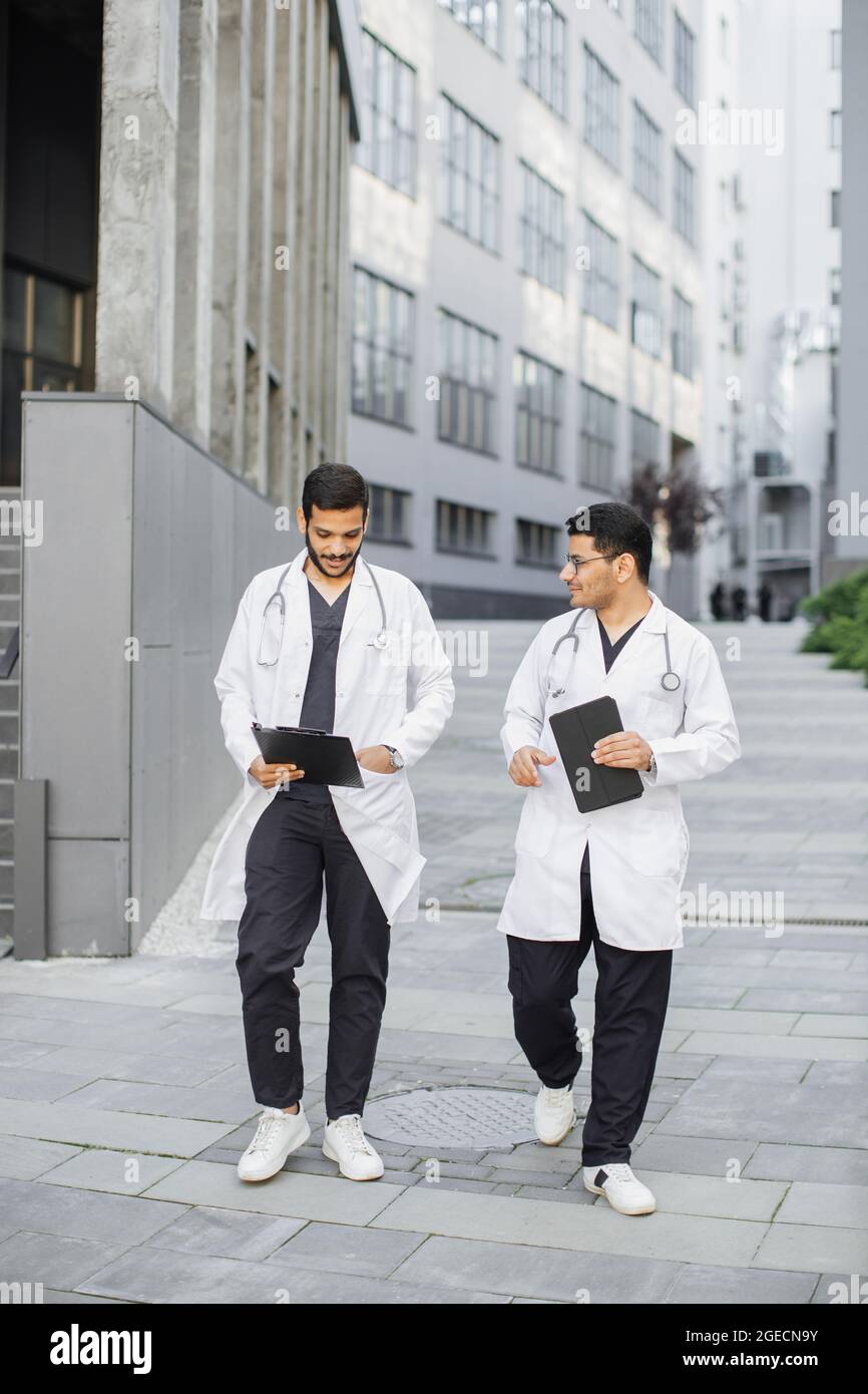 Two male Indian Arabian doctors in medical apparel, discussing patient's diagnosis, walking outside on the background of modern hospital Stock Photo