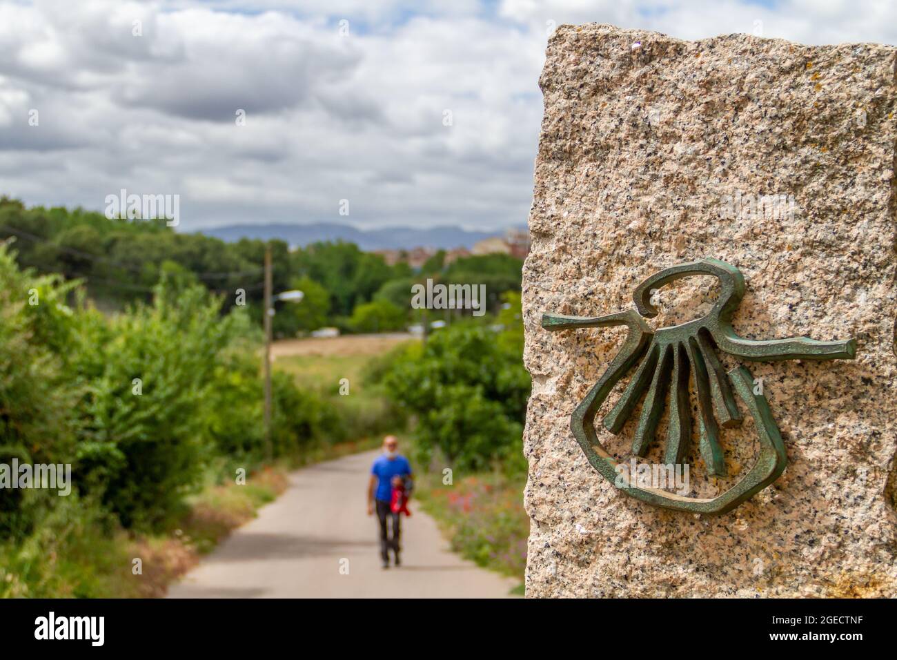 Pilgrim out of focus walks along the Camino de Santiago (Sant James Way) carrying a backpack next to a column on focus with the shell symbol of the Ca Stock Photo