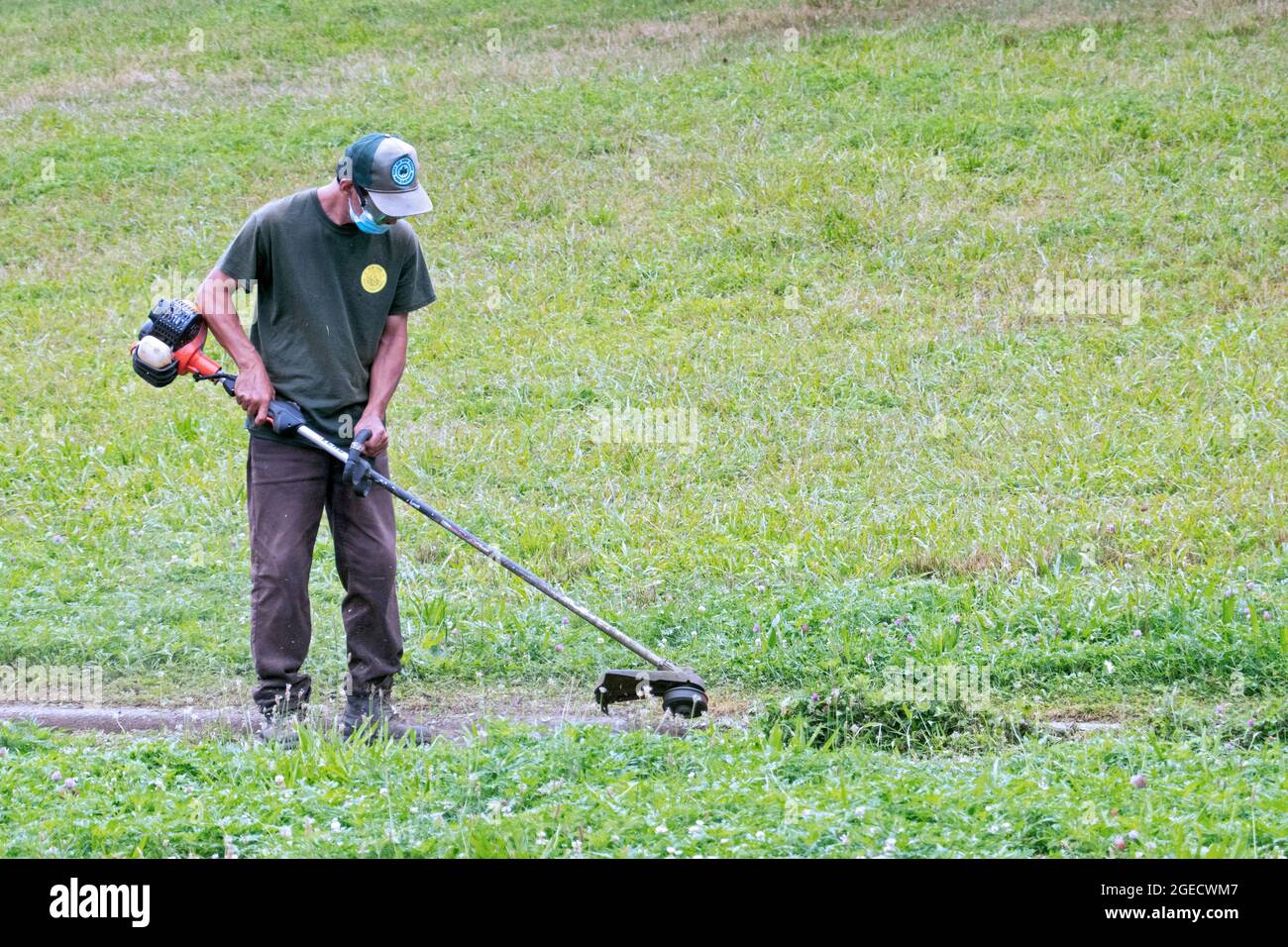 A New York City Parks Department worker uses a weed whacker to trim growth near a curb in a park in Queens, New York City. Stock Photo