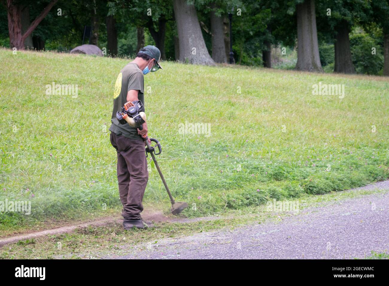 A New York City Parks Department worker uses a weed whacker to trim growth near a curb in a park in Queens, New York City. Stock Photo