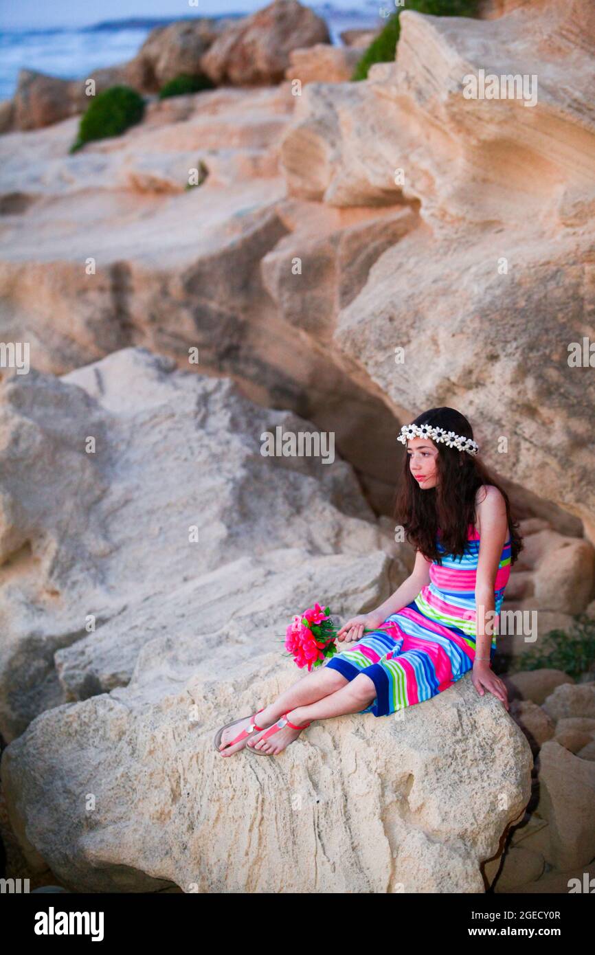 Preteen in colourful dress holds a flower bouquet while siting and waiting on the beach for the return of her long lost lover (or father) Stock Photo