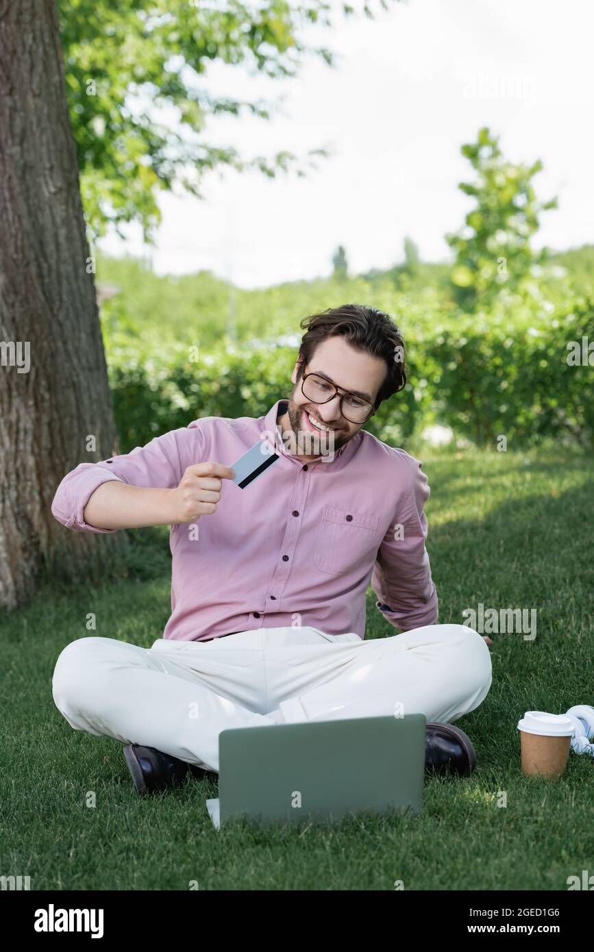 Cheerful businessman holding credit card during video call on laptop on grass Stock Photo