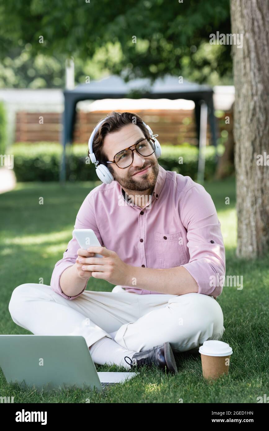 Dreamy businessman in headphones using gadgets near paper cup on grass Stock Photo