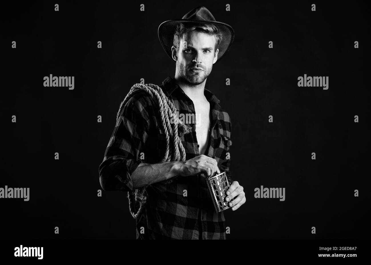 Western culture. Man wearing hat hold rope and flask. Lasso tool of American cowboy. Man handsome unshaven cowboy black background. Western life Stock Photo