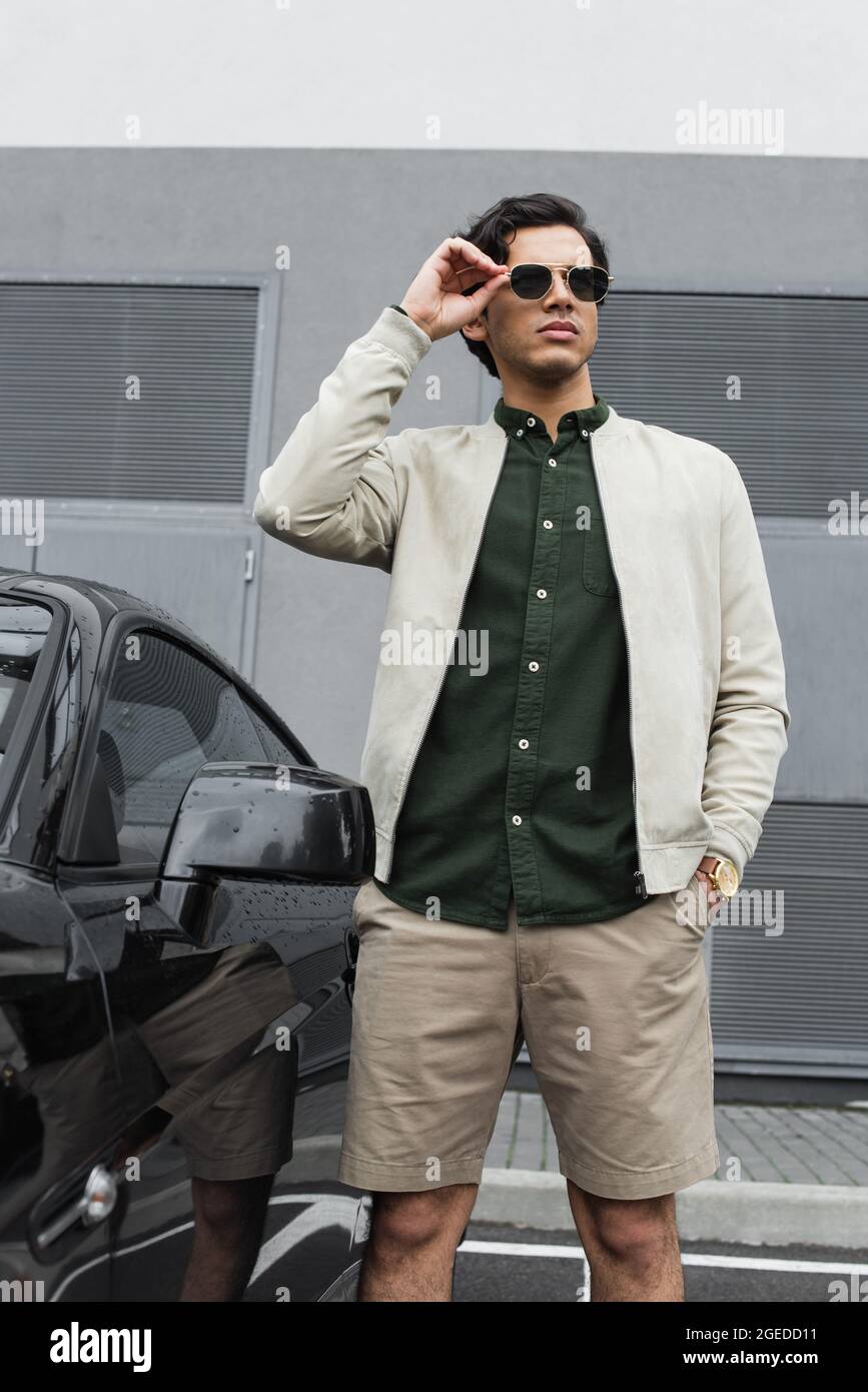 young man in stylish sunglasses and bomber jacket standing with hand in  pocket near modern car outside Stock Photo - Alamy