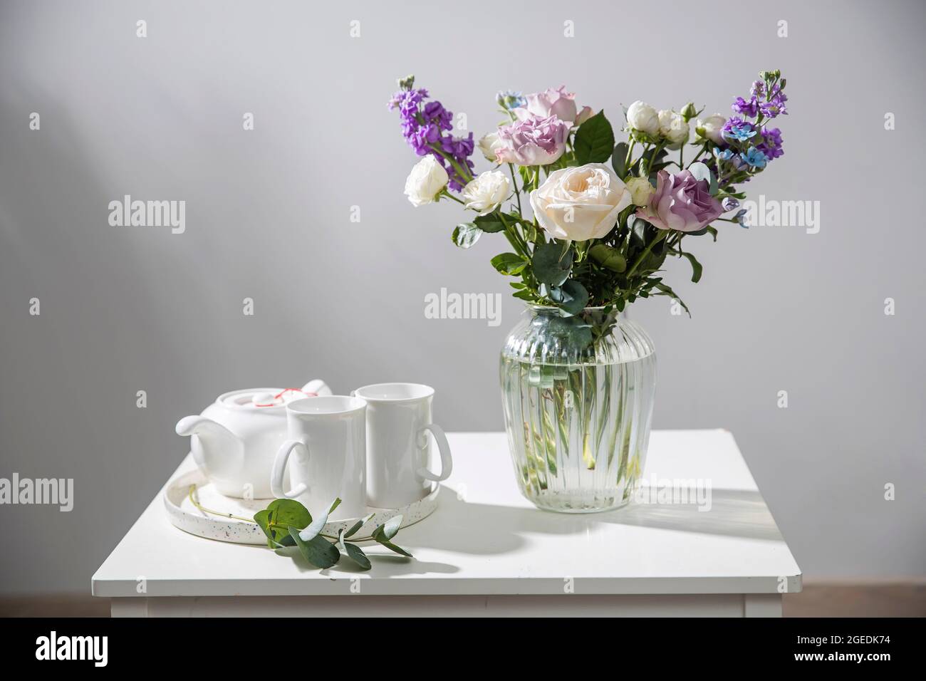 Bouquet of hackelia velutina, purple and white roses, small tea roses, matthiola incana and blue iris in glass vase is on the white coffee table. Grey Stock Photo