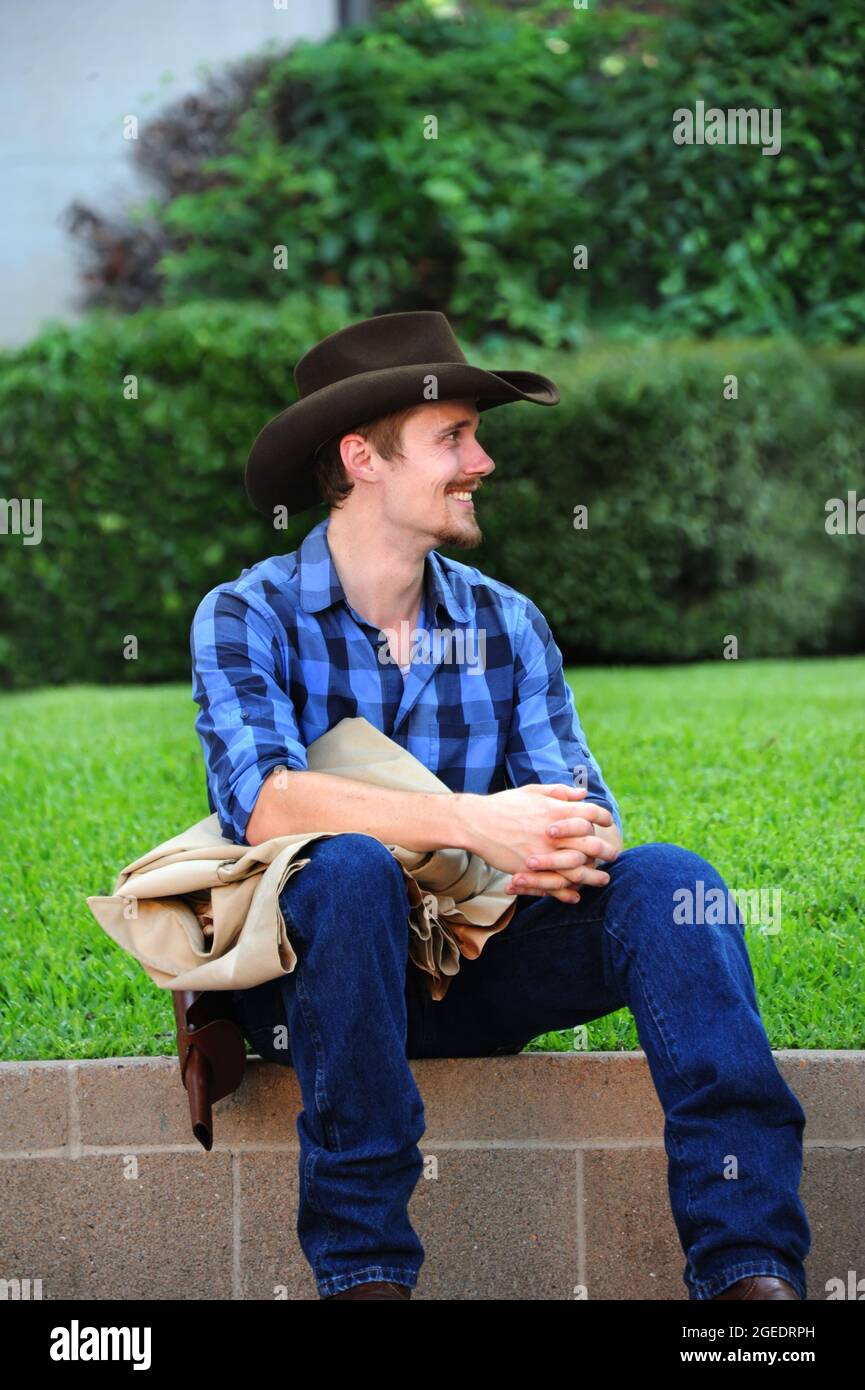 Young and handsome re-enactor sits waiting for his cue.  He is in a western theatre production and is wearing cowboy hat, jeans, gun belt and plaid sh Stock Photo
