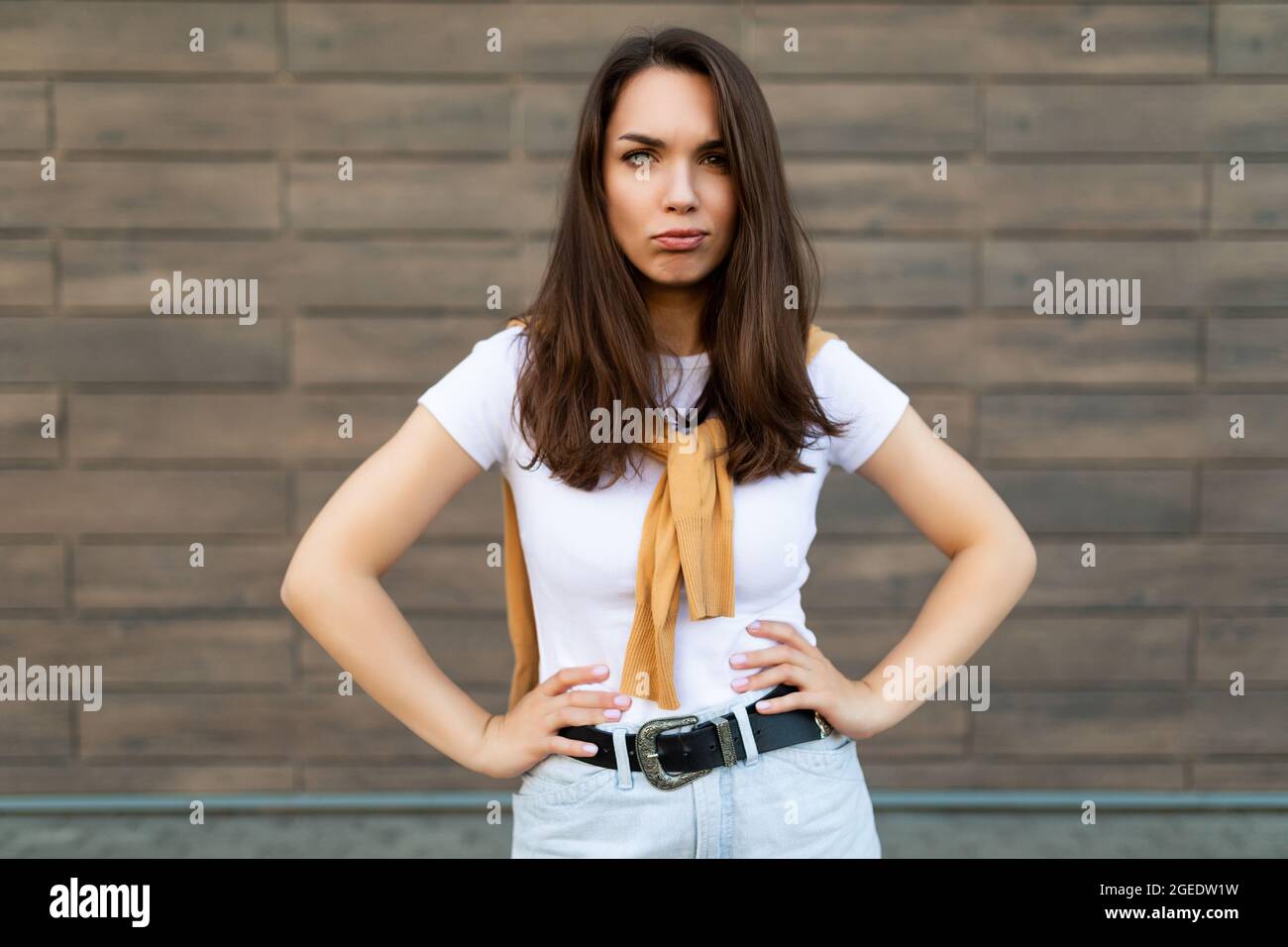 Portrait of touchy sad upset offended resentful young brunet woman wearing casual white t-shirt and jeans with yellow sweater poising near brown wall Stock Photo