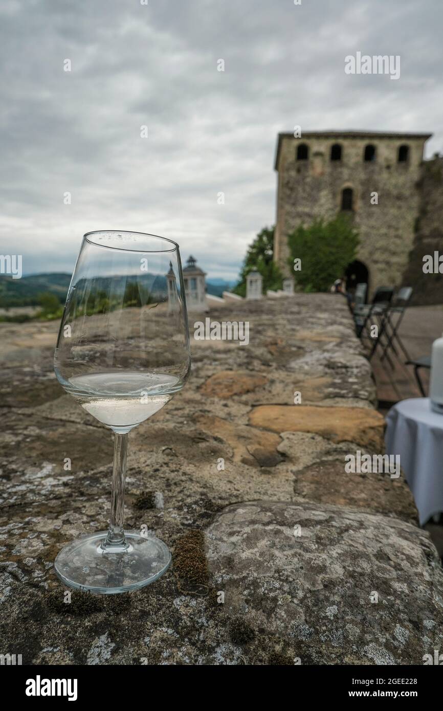 glass of white wine in restaurant across ancient castle wall, restaurant terrace and mountains view. Langhirano, Italy. Travel Stock Photo