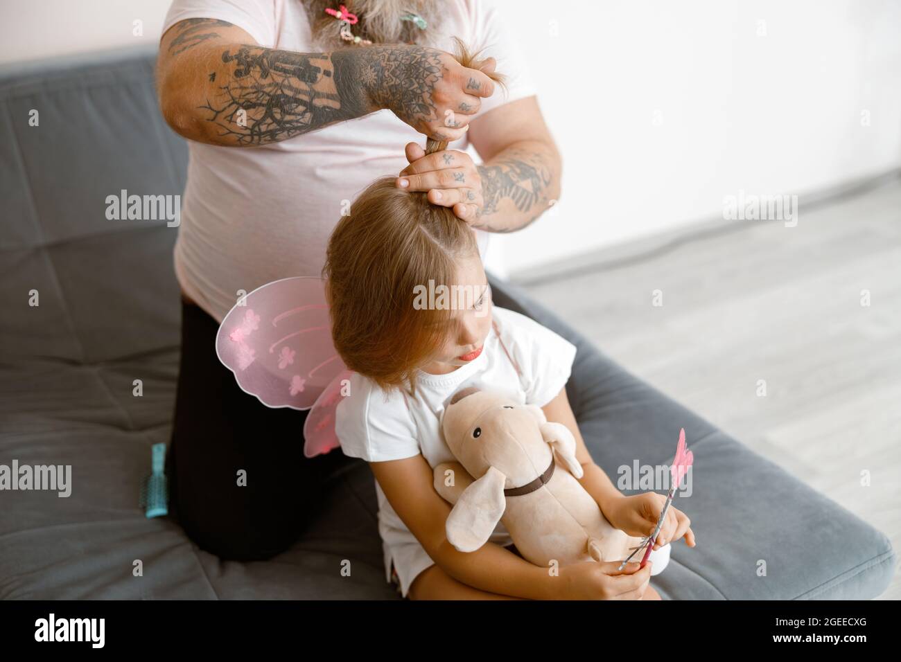 Father with tattoos makes ponytail to little girl wearing fairy suit in room Stock Photo