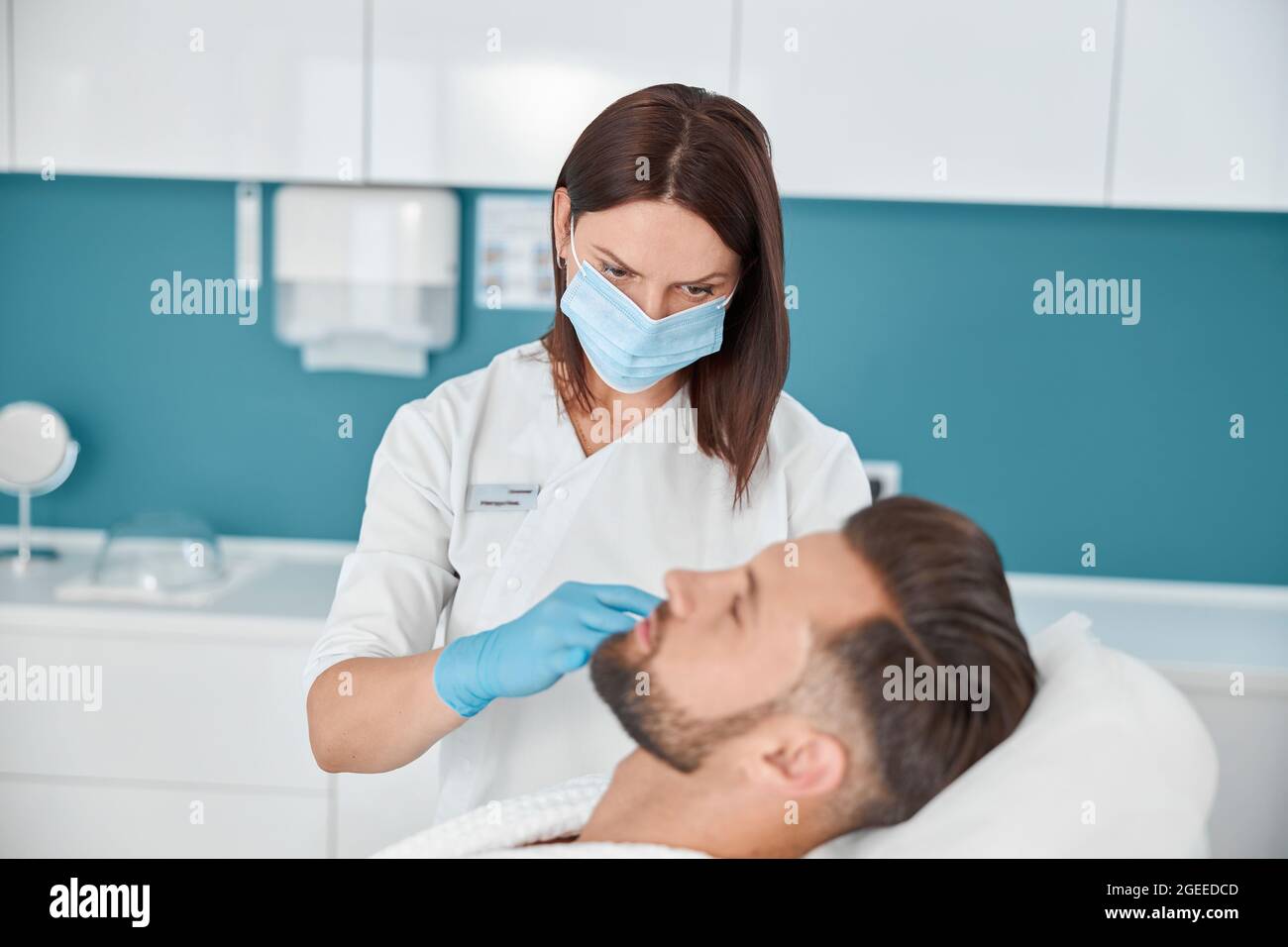 Woman with mask does injection to middle aged man at nasolabial fold filler procedure in clinic Stock Photo