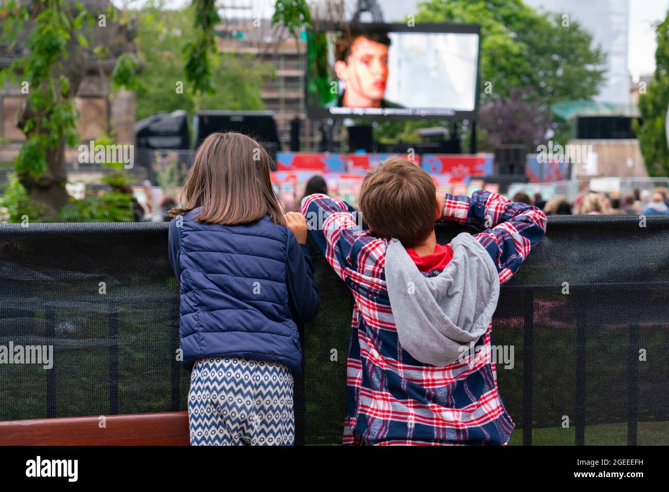 Edinburgh, Scotland, UK. 19th August  2021. Audience watch an outdoor screening of the cult movie Ferris Bueller’s Day Off at the  Film Fest in the City outdoor cinema in St Andrew Square. This is one of the events taking place during the Edinburgh International Film Festival in the city. Pic; Young movie goers watch the screening  from over the fence.  Iain Masterton/Alamy Live news. Stock Photo