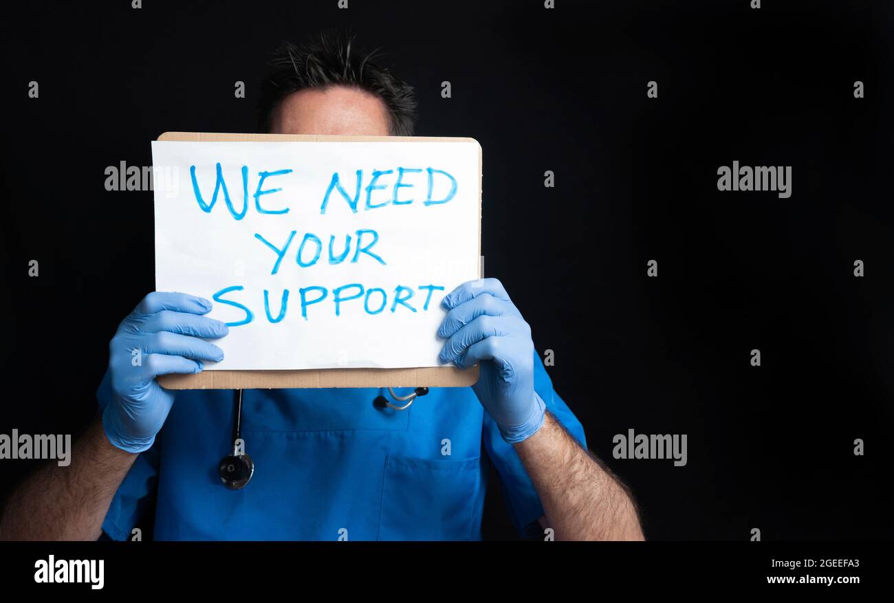 An unrecognizable, dark-haired doctor wearing gloves, a stethoscope and a medical uniform jacket holds a sign with his face covered, asking for suppor Stock Photo
