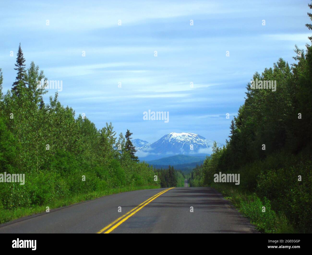 Alaskan highway continues toward snow capped mountain in the distance.  Car with headlights on traverses highway. Stock Photo