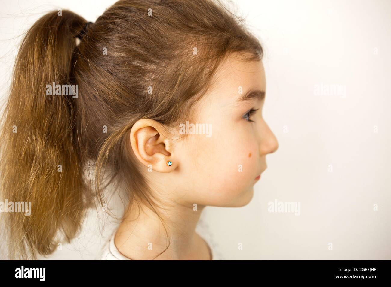 Ear piercing in a child - a girl shows an earring in her ear made of a medical alloy. White background, portrait of a girl with a mole on his cheek in Stock Photo