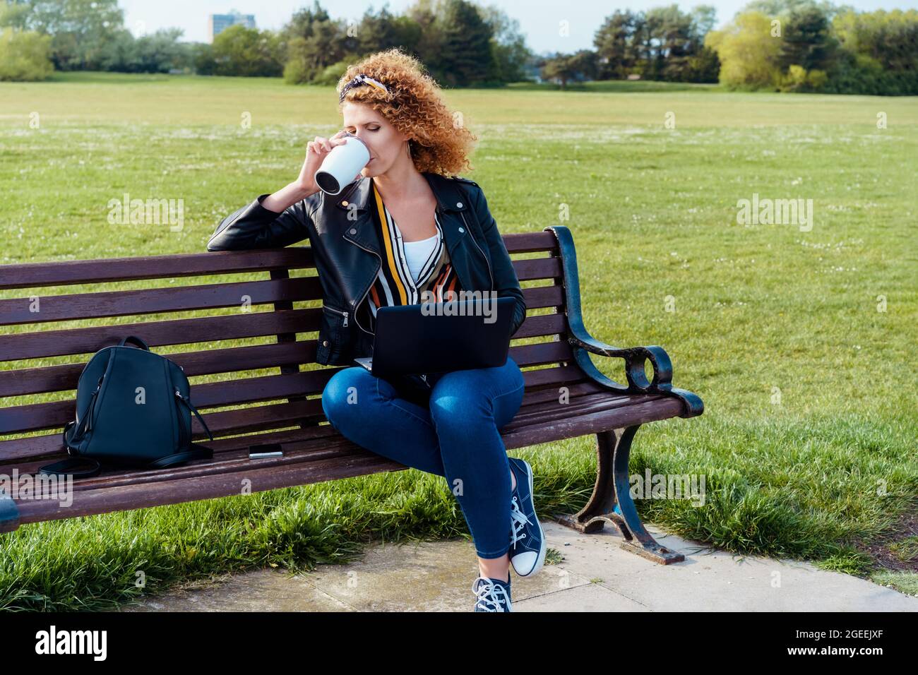 A woman in casual clothes drinking coffee from reusable cup and using a laptop outdoors sitting on the bench in the public park. Remote work Stock Photo