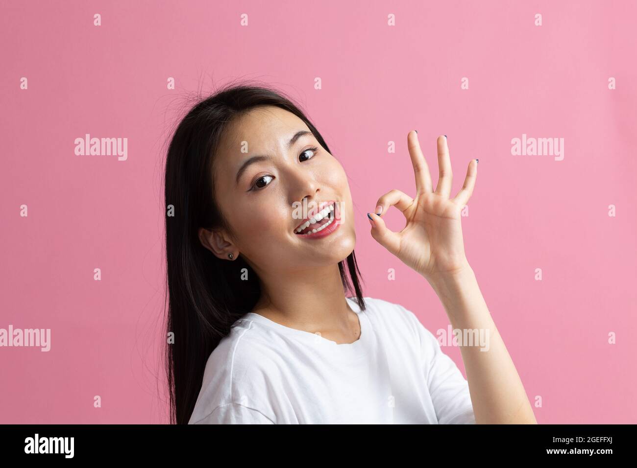 Smiling asian girl showing Ok gesture. Beautiful young woman with dark hair wearing white t-shirt. Female looking at camera isolated on pink background. Studio shoot Stock Photo