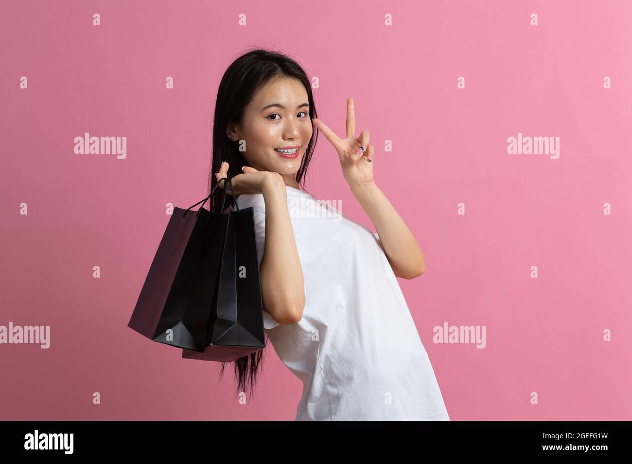 Shopping asian happy woman holding shopping bags v gesture on pink background. Stock Photo