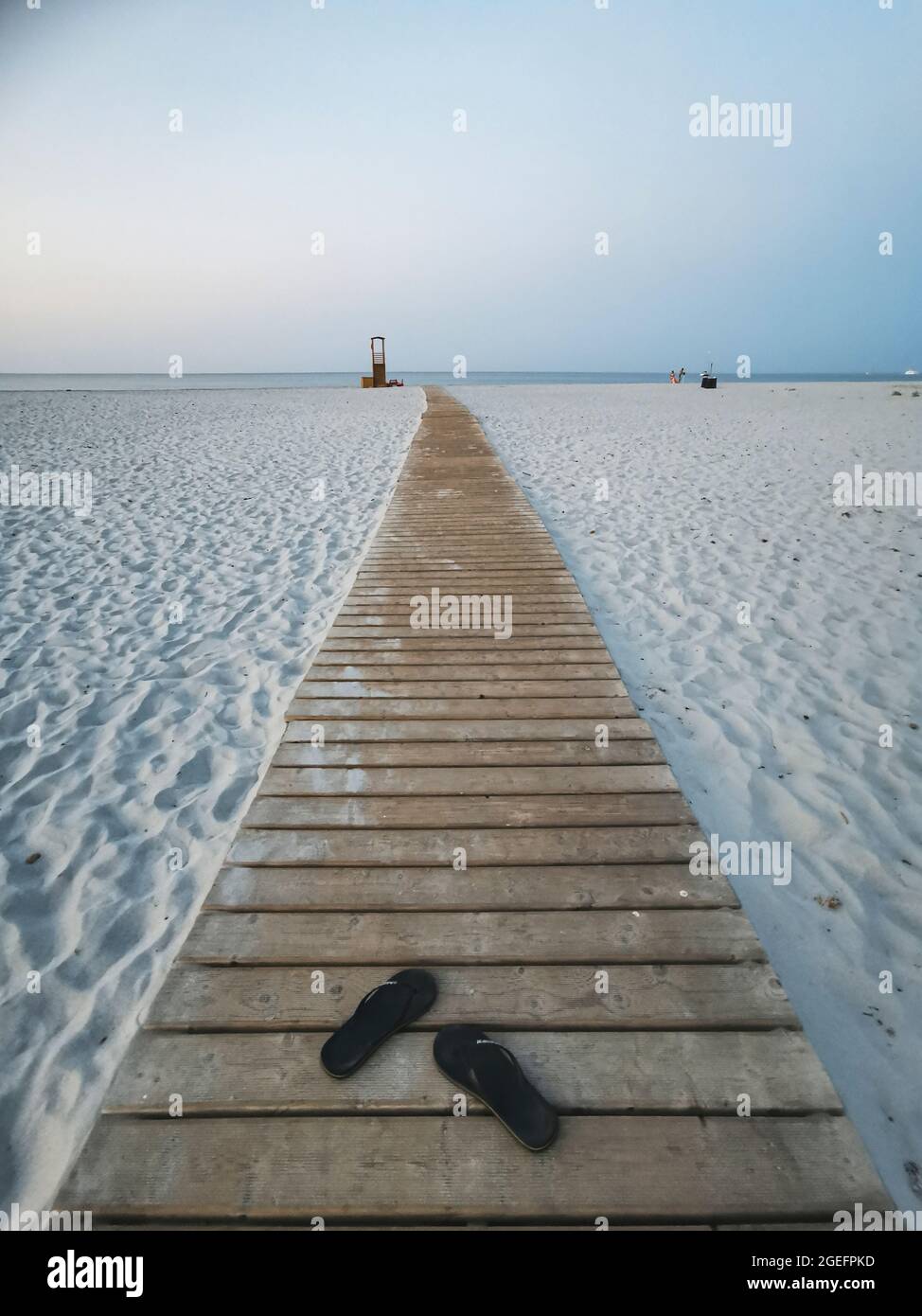 Long wooden path on the white beach sand to sea water with pair of sandals alone - concept of people and summer holiday vacation - ocean and sky view Stock Photo