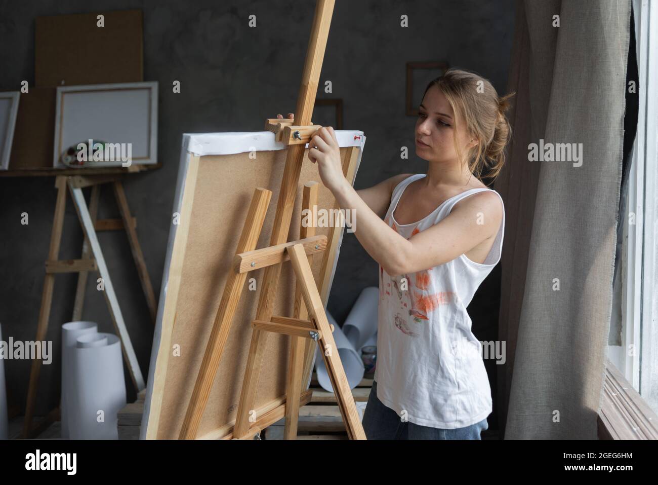 Portrait of a woman artist in an art studio stands near the easel and fixes the canvas. Stock Photo