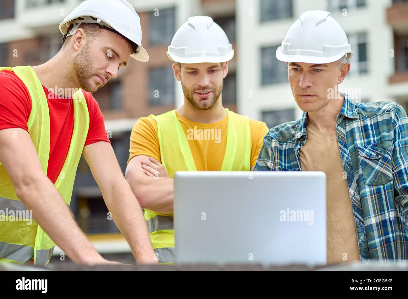 Three concentrated men in protective helmets standing before a laptop Stock Photo