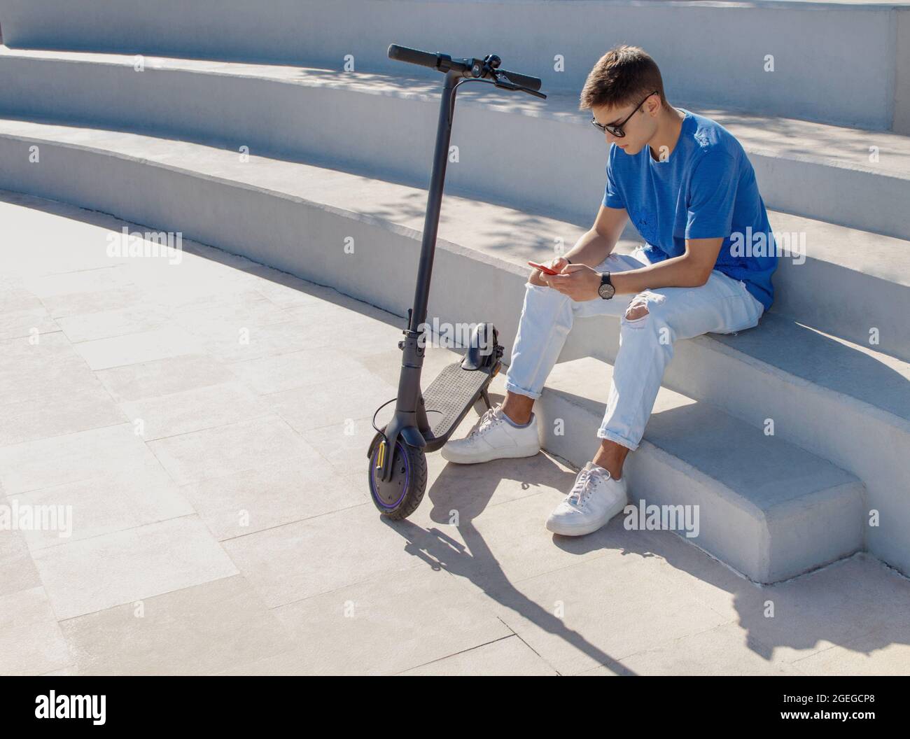 Young caucasian man in blue t-shirt sitting outdoor on stone stairs near electric scooter and using his phone. Electro mobility concept Stock Photo