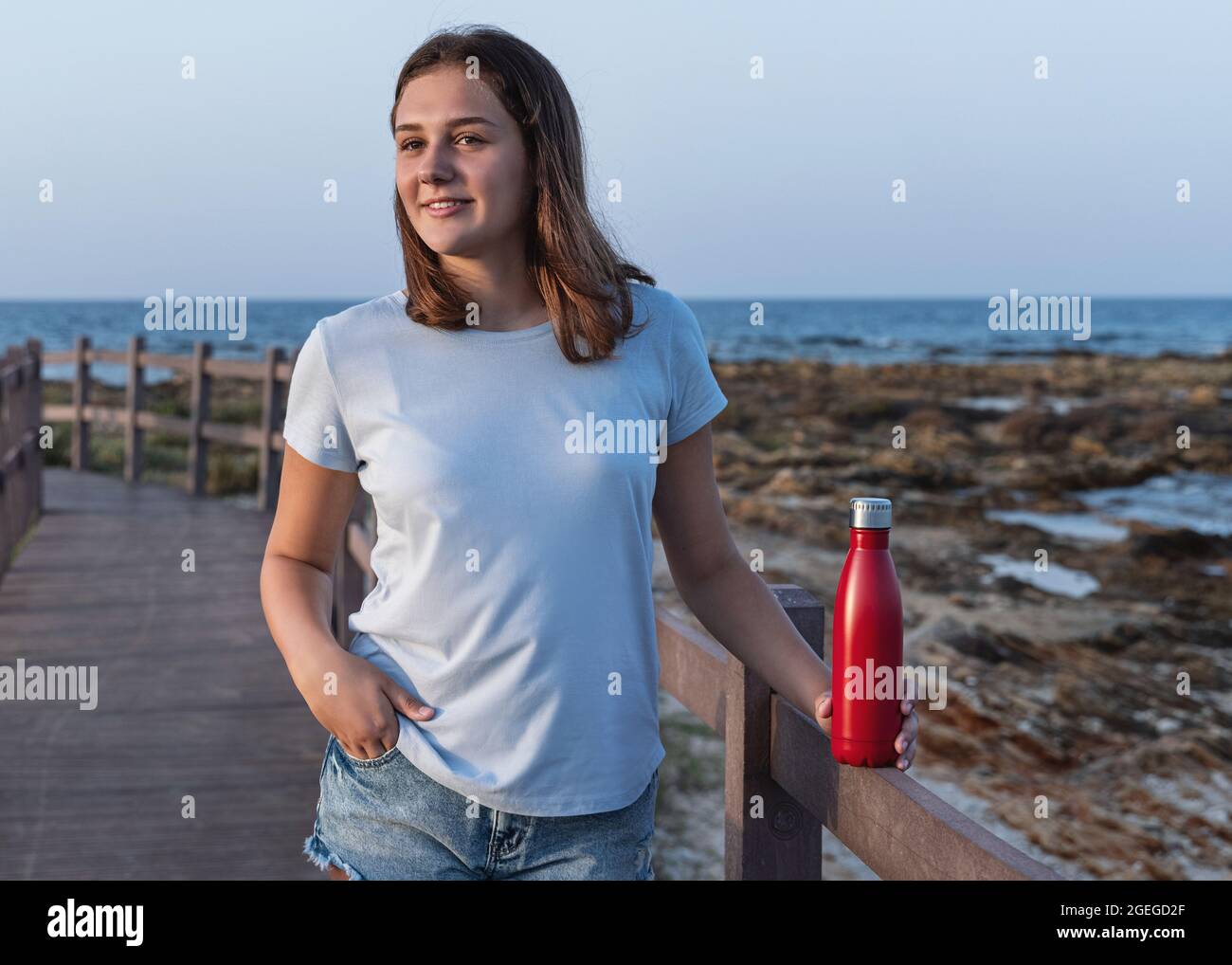 Teen girl in blue t-shirt standing and holding red reusable water bottle in hands by the sea, three quarter length shot. Bottle and t-shirt mockup Stock Photo