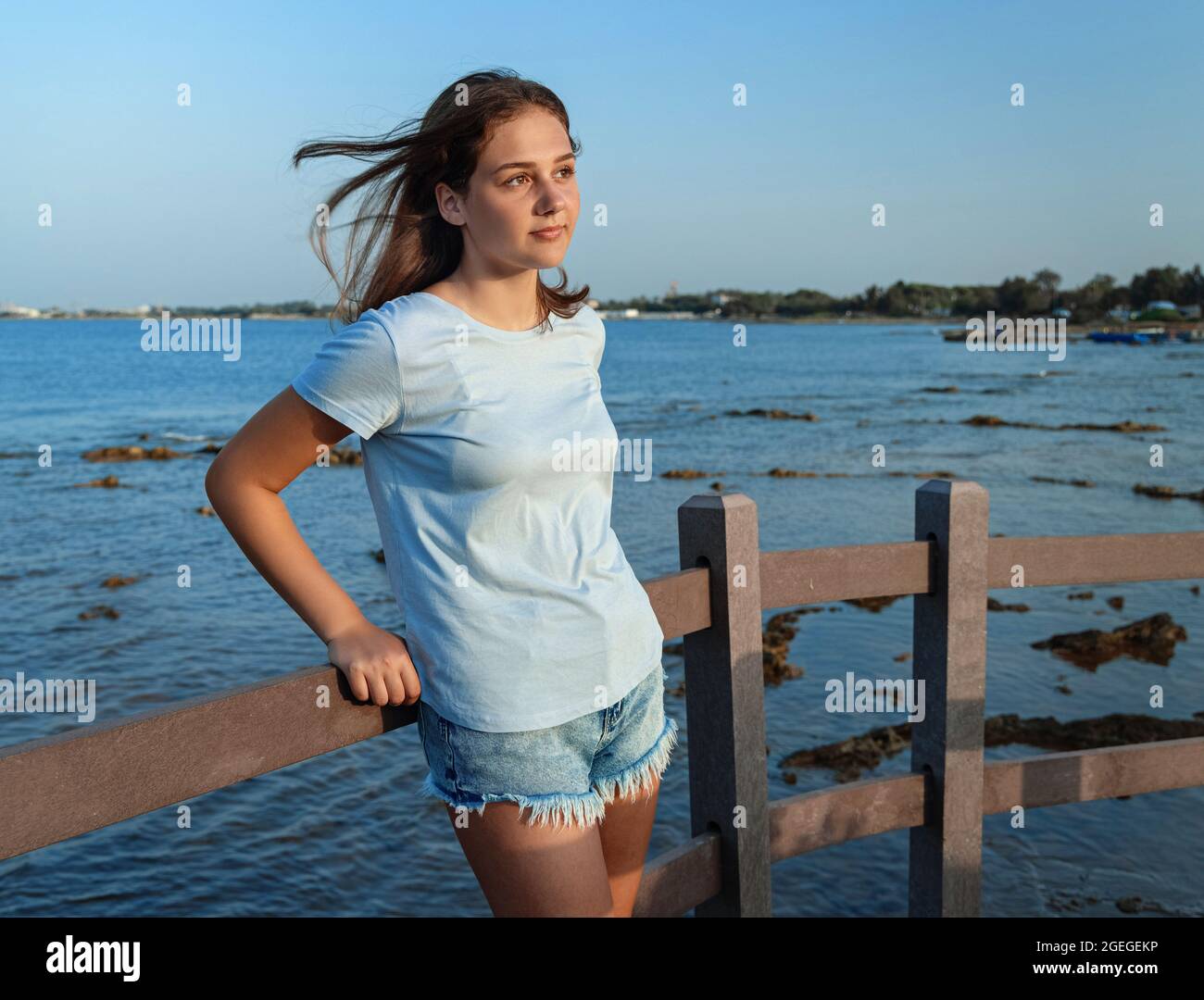 Teen girl standing on on wooden bridge by the sea at sunset. Teenager wearing blue t-shirt and jeans shorts looking aside, three quarter length shot. Stock Photo