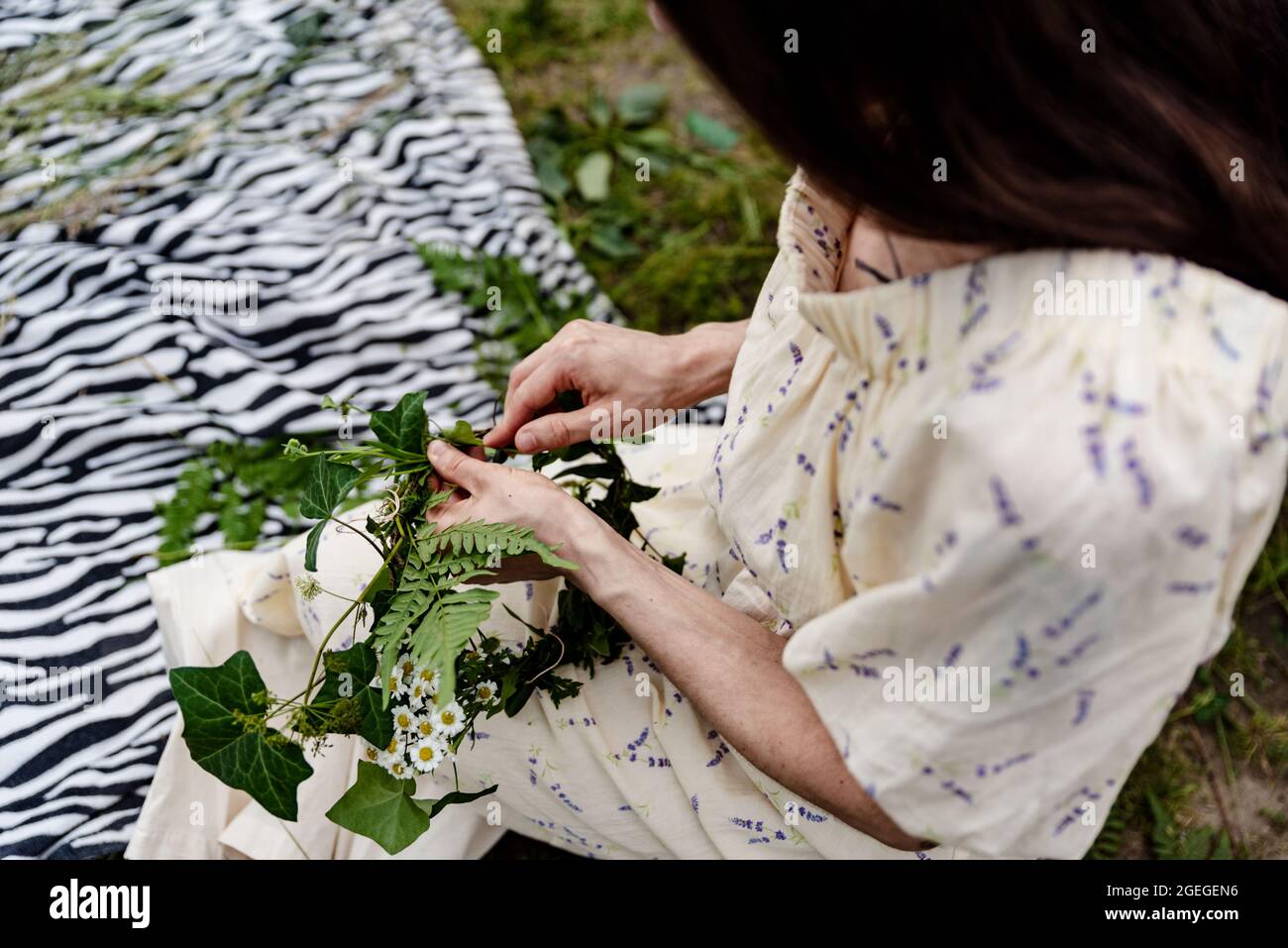 Woman making head wreath with wild flowers. Close up of hands weaving a wreath with green leaves and flowers on midsummer day Stock Photo