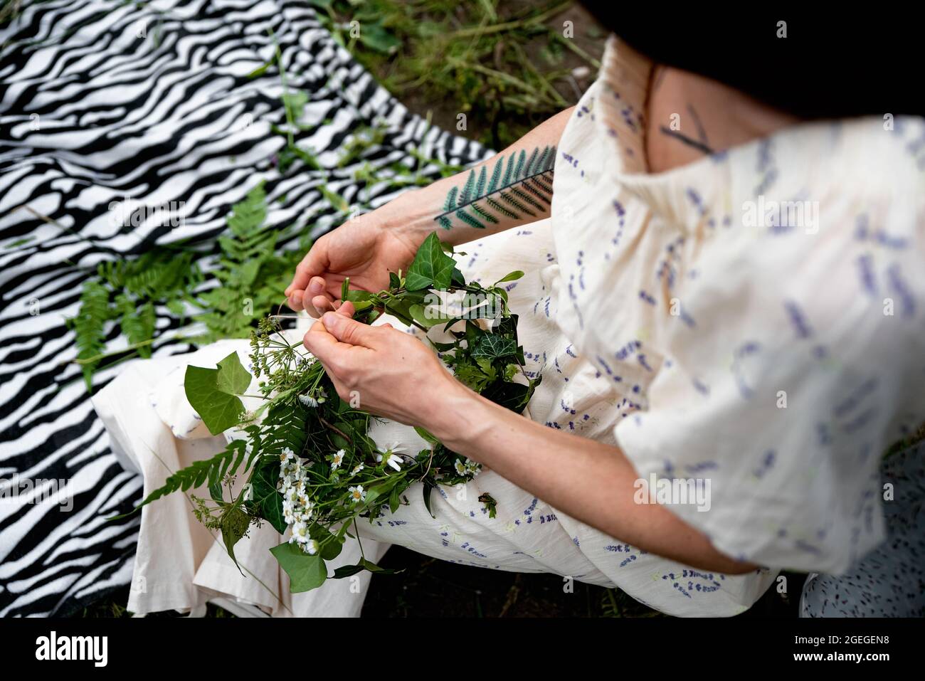 Woman making head wreath with wild flowers. Close up of hands weaving a wreath with green leaves and flowers Stock Photo