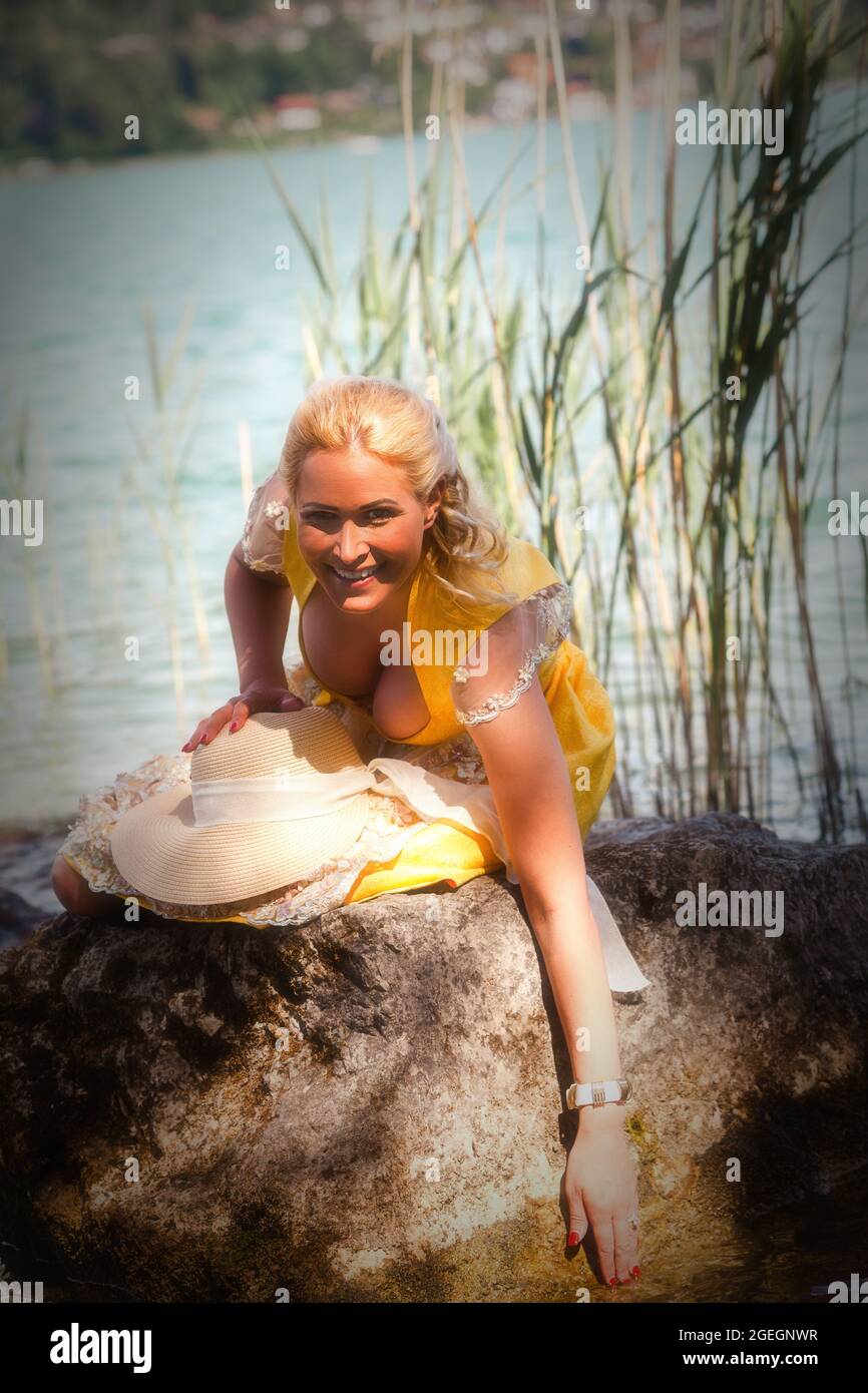 Elegant woman with dirndl sits smiling on a stone in the lake and holds one hand in the water. Stock Photo