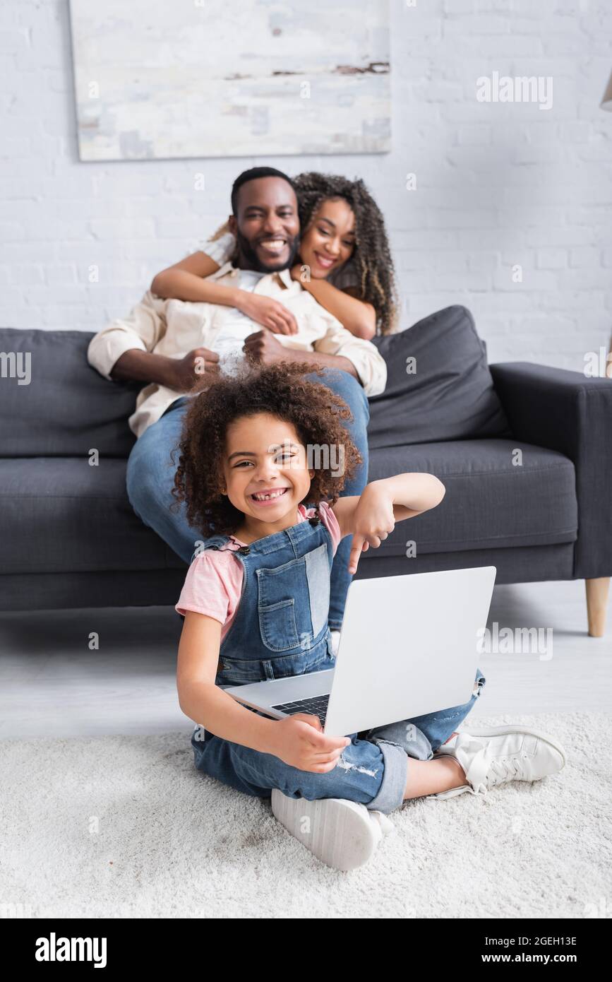 happy african american girl pointing at laptop while sitting on floor ...