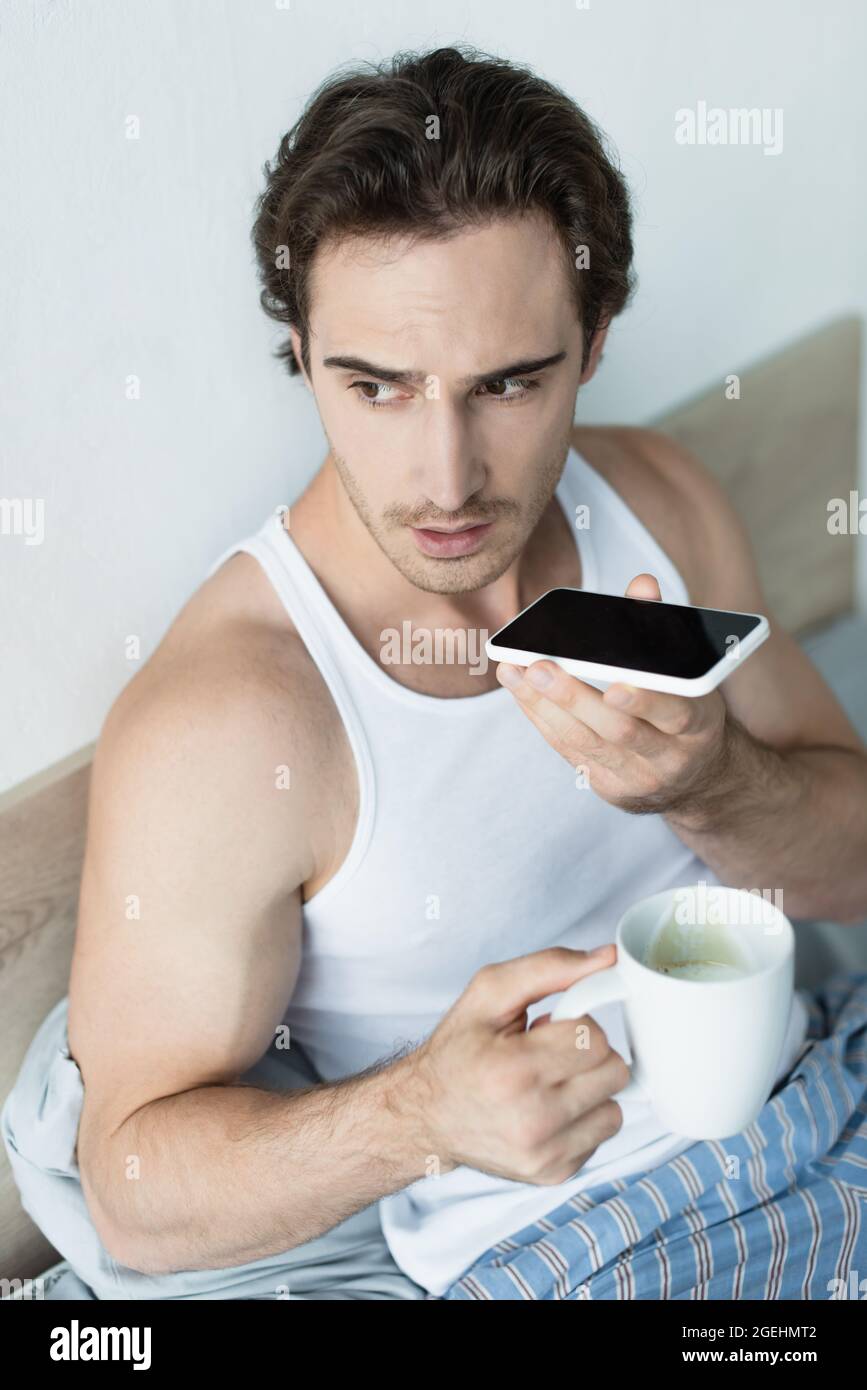 brunette man holding cup of coffee and sending voice message on smartphone in bedroom Stock Photo