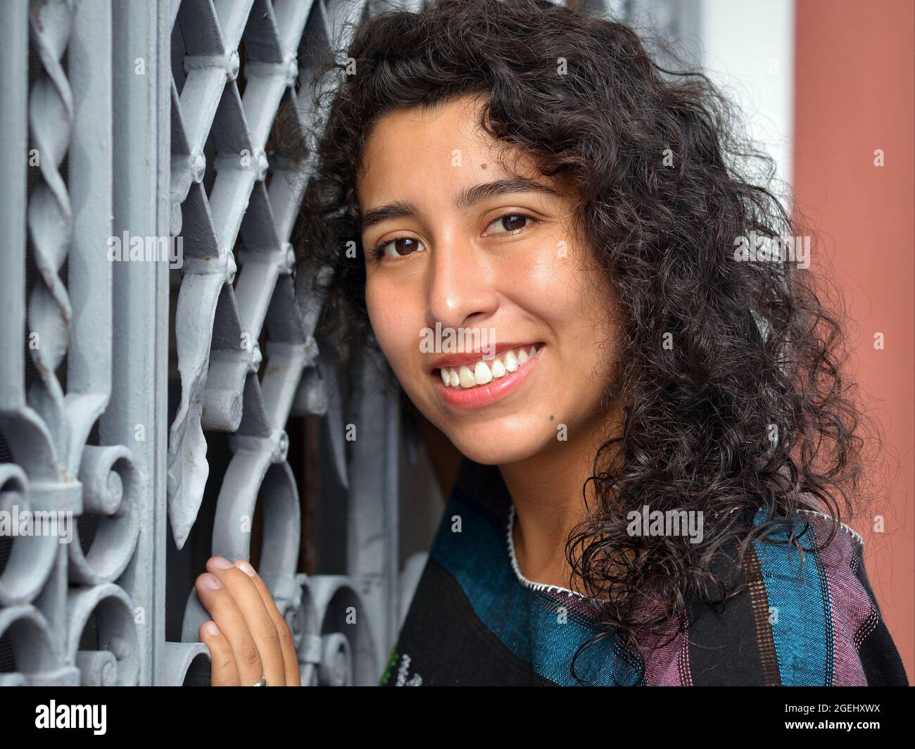 Beautiful positive emotional young Mexican Latina Hispanic woman with long black curls poses in front of a white security steel window grill. Stock Photo