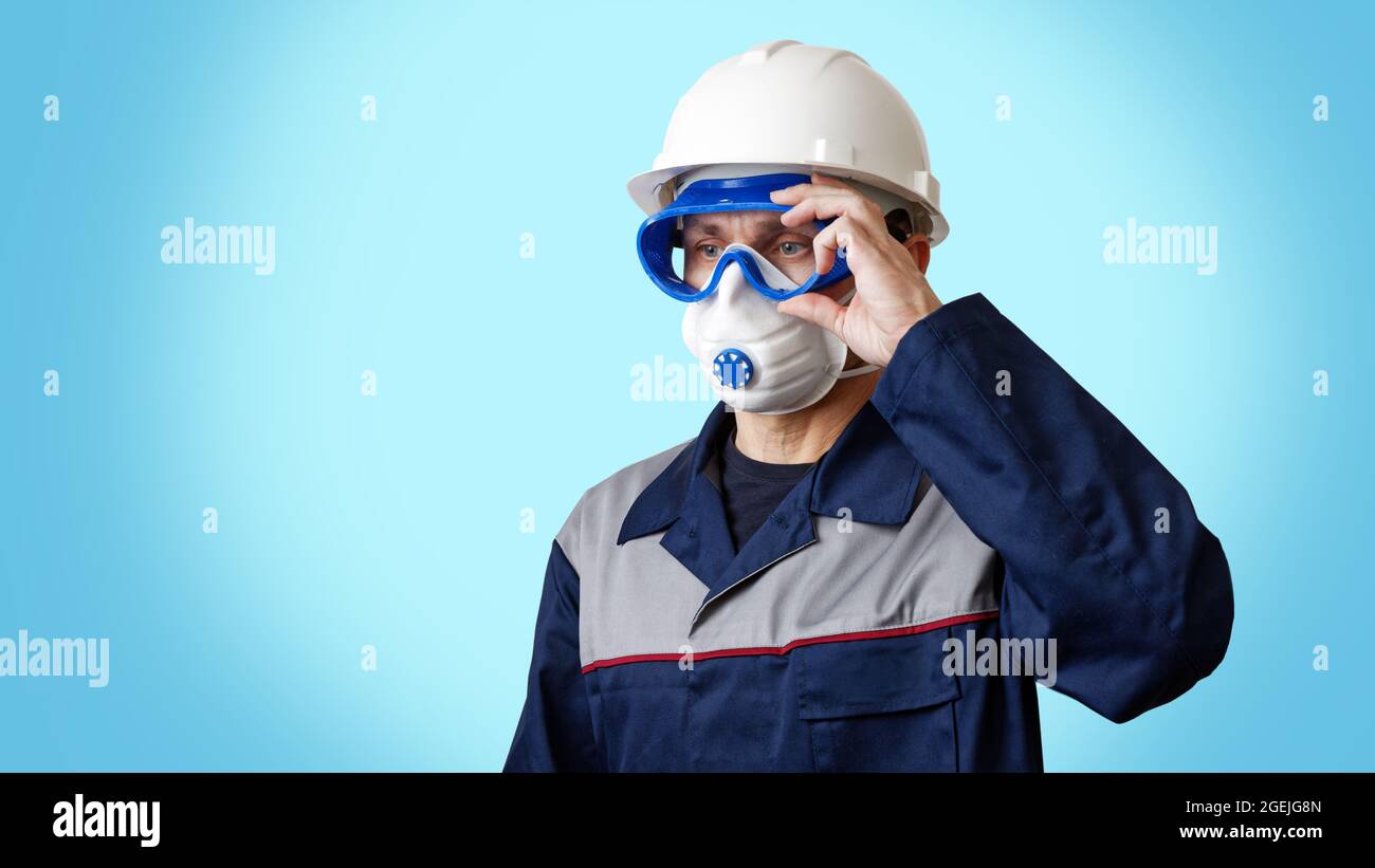 Portrait of a man in a work clothes, white construction helmet, respirator and protective glasses, isolated on blue. Copyspace. Stock Photo