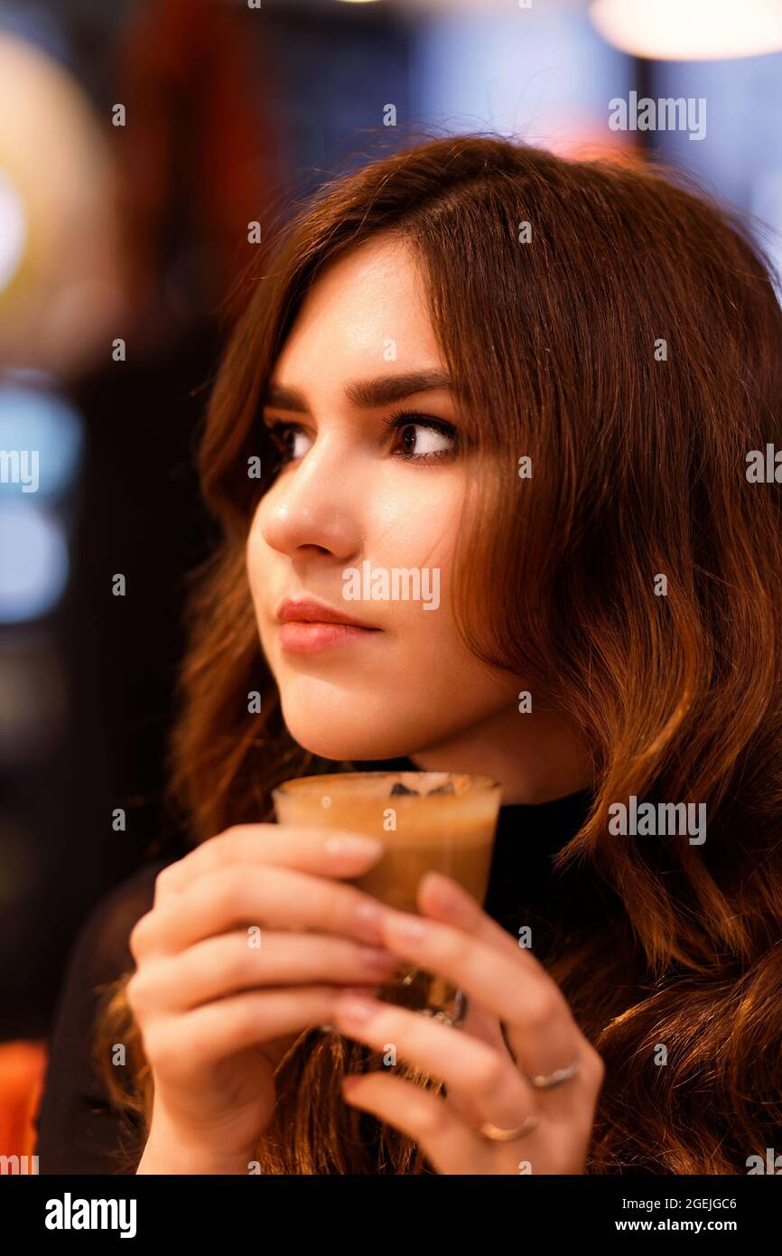 Portrait of a young girl holding a cup of coffee flat white in her hand and smiling while looking directly into the camera. Shallow focus and blurred Stock Photo